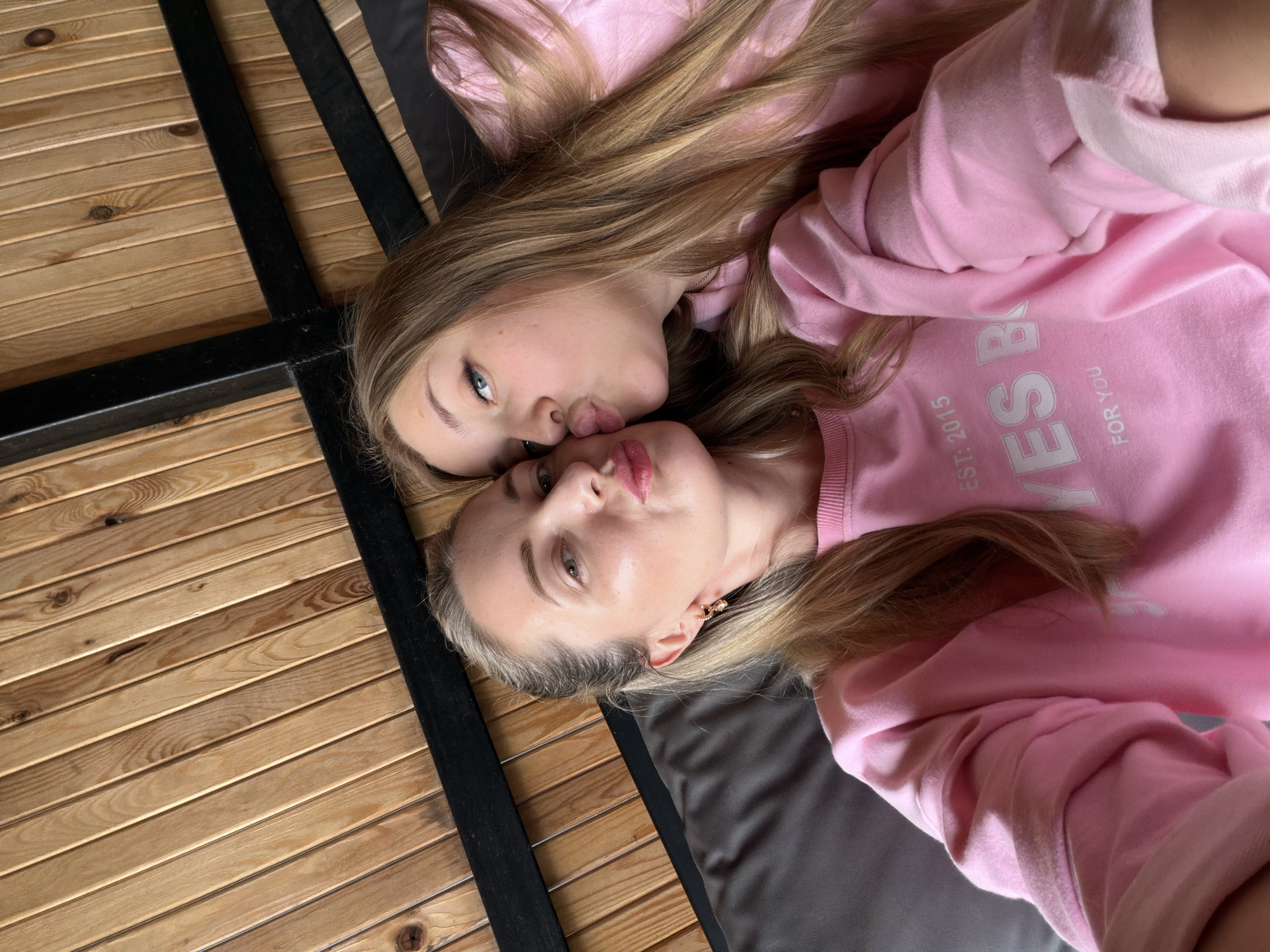 In this close-up selfie, two young women, likely mother and daughter, are captured from the chest up, lying down. The woman on the left, with blonde hair pulled back, is looking directly at the camera with pursed lips, while the woman on the right, also with blonde hair, leans in to kiss her cheek, her eyes looking towards the camera. Both are wearing matching pink sweatshirts. The sweatshirt on the left has the text "EST: 2015 YES B FOR YOU" in white lettering. They are lying on a dark grey surface, possibly a bed or couch, against a backdrop of light brown wooden planks arranged vertically. Black metal beams run horizontally and diagonally across the top portion of the image, suggesting a bunk bed frame or similar structure. The lighting is soft, illuminating their faces and clothing. The overall impression is one of affection and intimacy. The location is not specified in the image, nor is it possible to determine the city of Семилуки, Russia from the visual information provided.