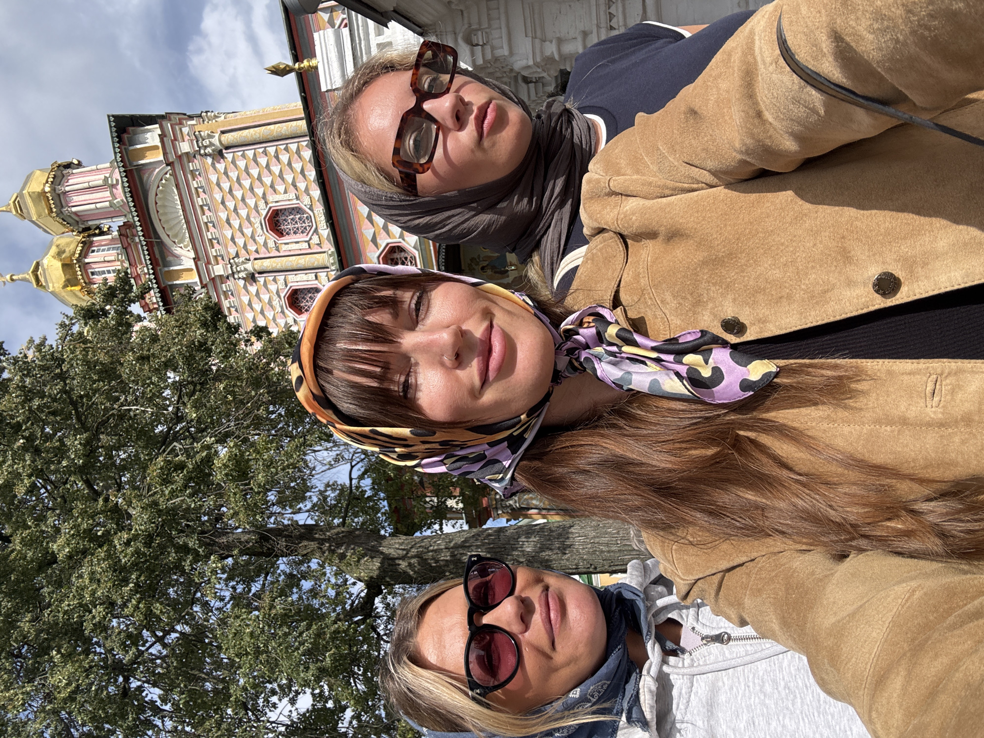 Three women are taking a selfie in front of a vibrant, ornate building. The woman in the center is wearing a brown suede jacket and a colorful headscarf with a leopard print pattern. Her hair is long and brown. To her left, a woman with blonde hair wears a gray hoodie and a blue bandana on her head, along with sunglasses with pink lenses. To the right of the central figure, a blonde woman wears a dark-colored headscarf and large, brown-framed glasses, a dark t-shirt is visible.
The building behind them is elaborately decorated with colorful geometric patterns in shades of pink, yellow, and white. It features multiple domes, with the two prominent ones in the background covered in gold leaf. The architecture suggests a Russian Orthodox church. The scene appears to be outdoors, with a tree on the left providing some shade and a hint of blue sky with scattered clouds above. The overall impression is a tourist snapshot taken at a significant religious or historical site in Sergiyev Posad, Russia, likely the Trinity Lavra of Saint Sergius, given the distinct architecture of the building.