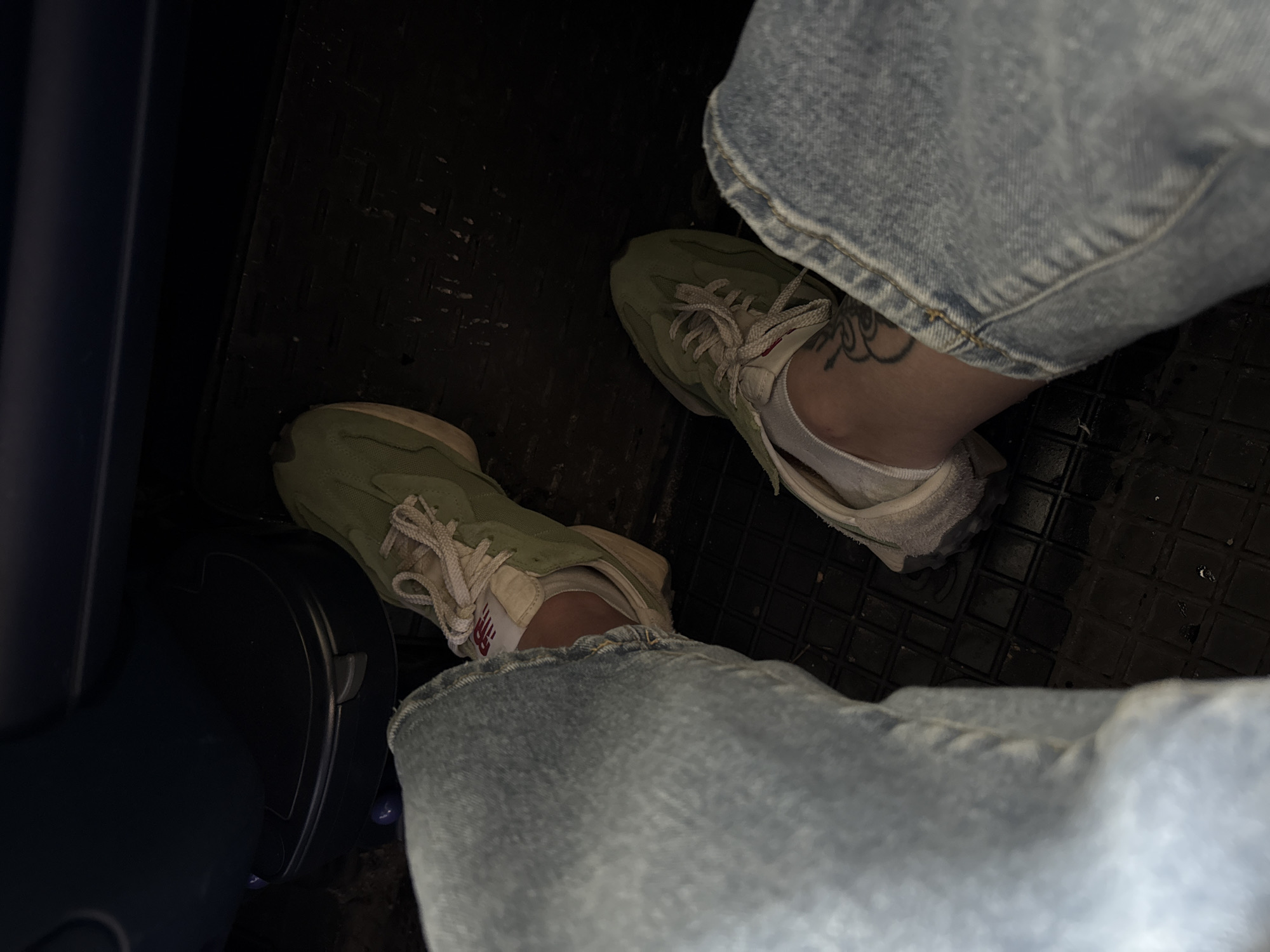 The image displays a close-up view of a person's legs and feet, wearing light-wash denim jeans and olive-green sneakers. The sneakers have white laces and a cream-colored sole. The person is seated, with their legs bent at the knees. The background suggests an indoor environment, possibly a vehicle or a room with dark flooring. A dark blue object is visible in the upper portion of the image, possibly a seat. A tattoo is visible on the ankle of the person's right leg. The lighting is dim, creating shadows and a slightly grainy texture in the image.