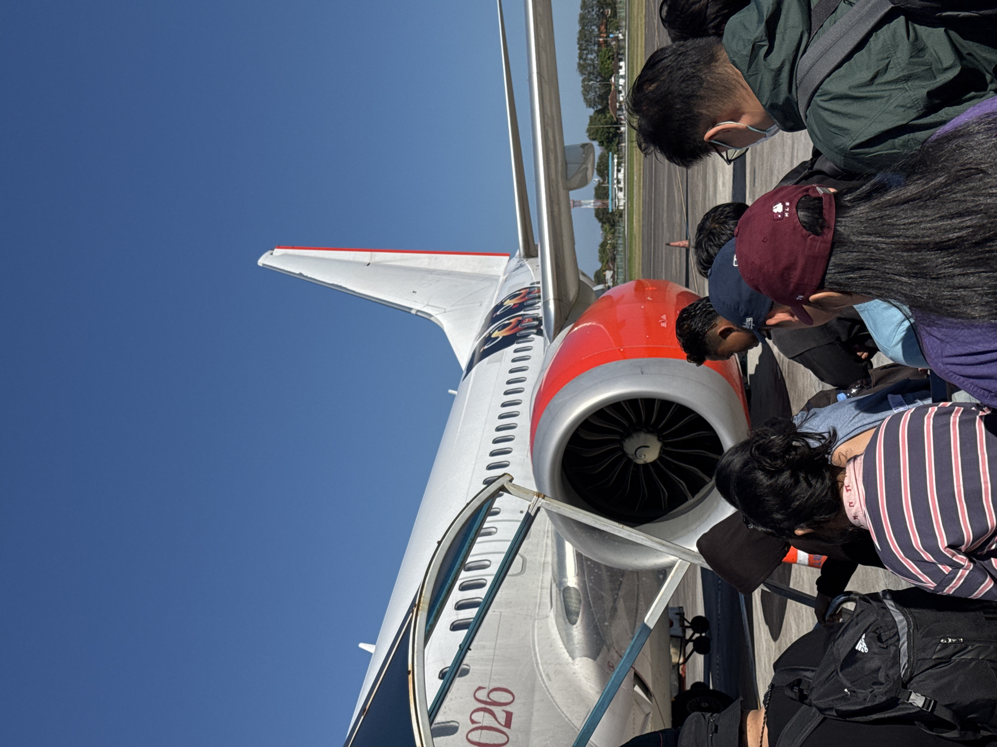 In Kuta, Indonesia, a group of passengers, some wearing face masks and hats, are lined up on an airport tarmac. They are waiting to board a white airplane with red accents, identified by the registration number "026". The plane's engine is visible, and the tail fin features red and white stripes. The sky is clear and blue, and the ground is paved. In the background, distant buildings and trees are visible. The scene appears to be a typical airport boarding process under sunny weather.