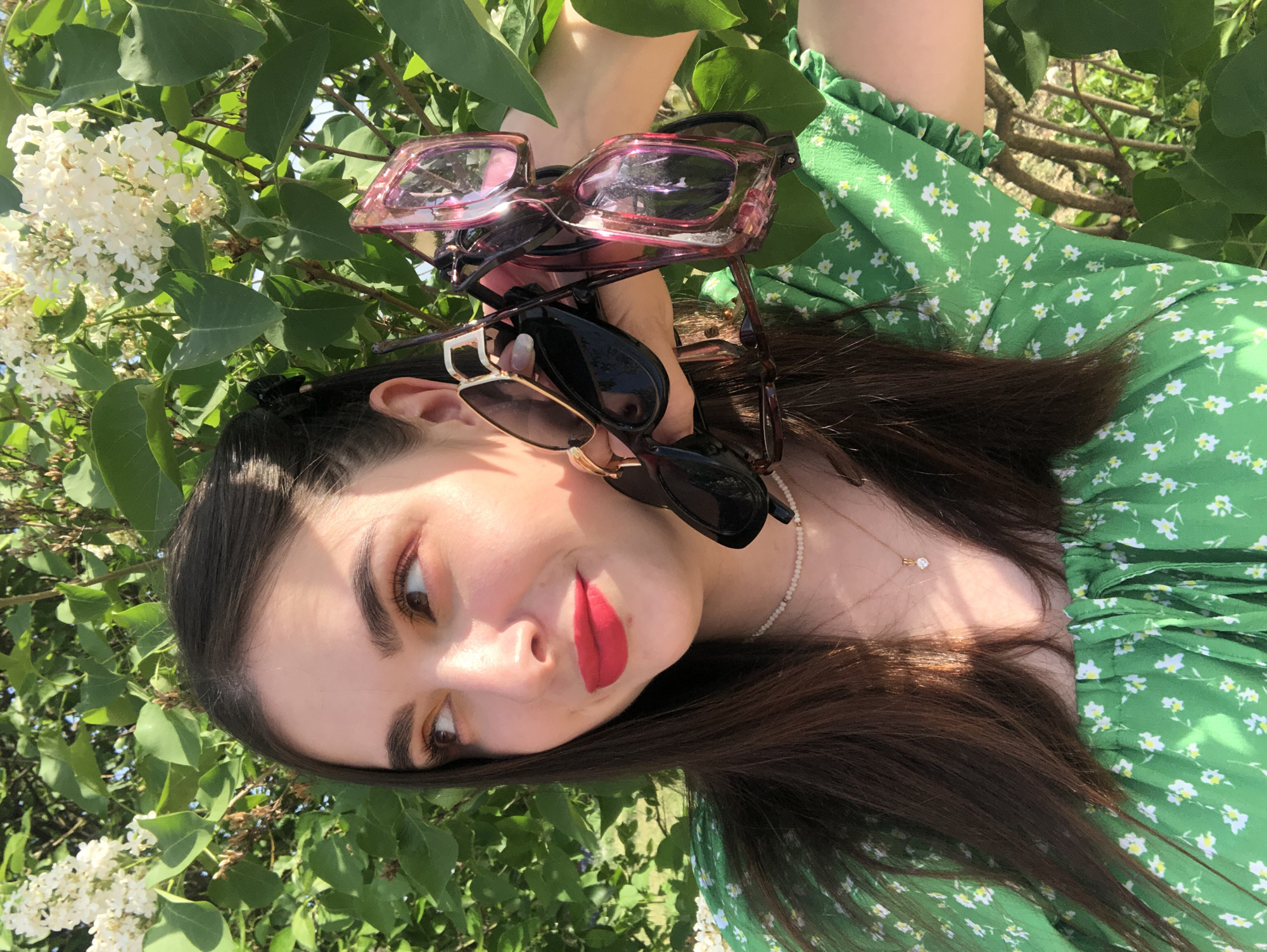 A woman poses outdoors with lilac blossoms in the background. She has long, dark hair and is wearing a green dress with small white floral patterns. She is holding multiple pairs of sunglasses in her hand. She has fair skin, brown eyes, and red lipstick. She wears two necklaces, one white and one gold. The location is Noviy, Russia.