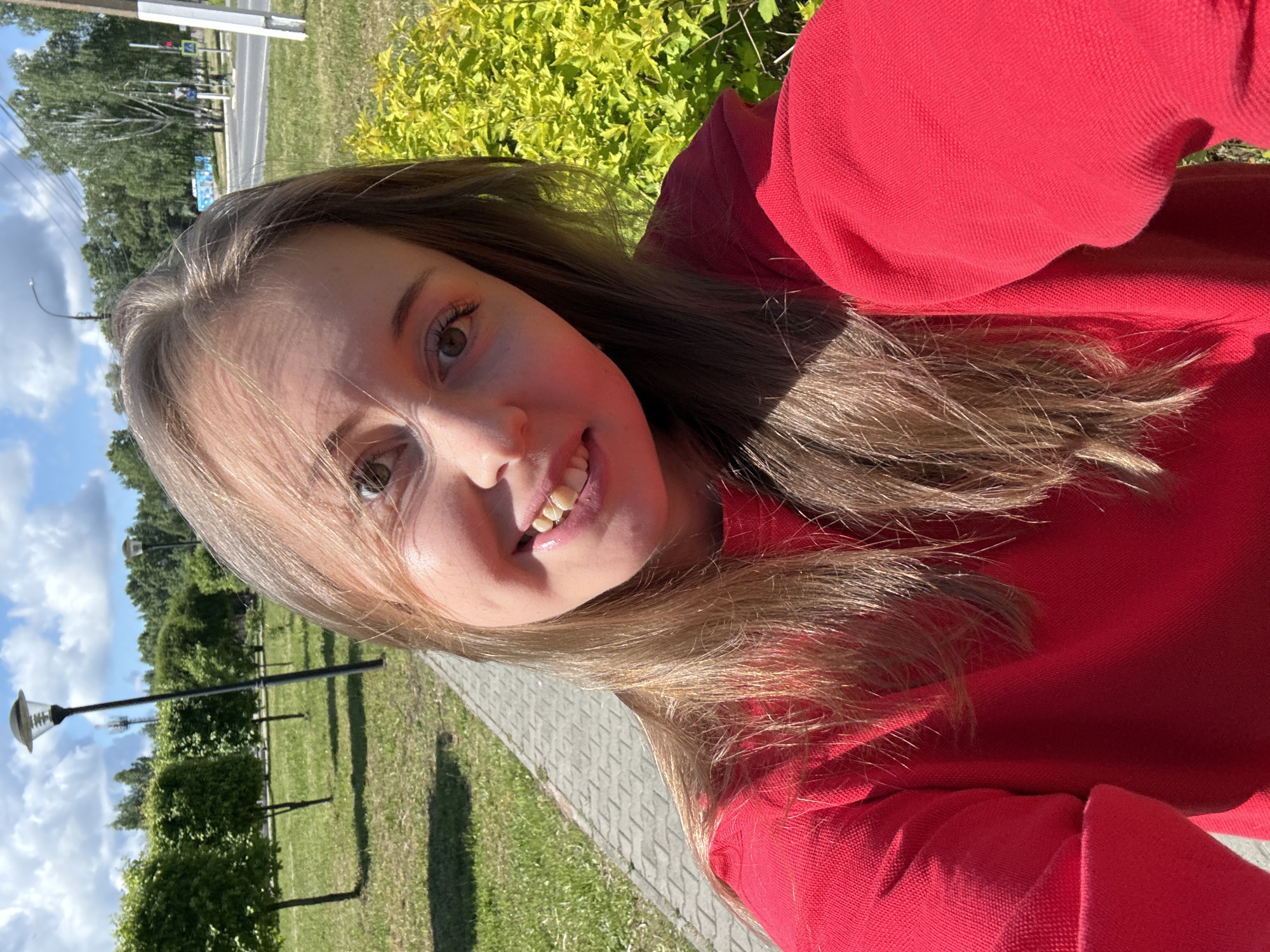 A young woman with long, light brown hair is posing for a selfie outdoors. She is wearing a red long-sleeved shirt or sweater. She has a slight smile showing her teeth.
Behind her, there is a paved walkway with grass and trees on either side, as well as streetlights. In the distance, across a road, more greenery and possibly buildings can be seen. The sky is blue with some clouds. The environment suggests a park or a landscaped area in a city.