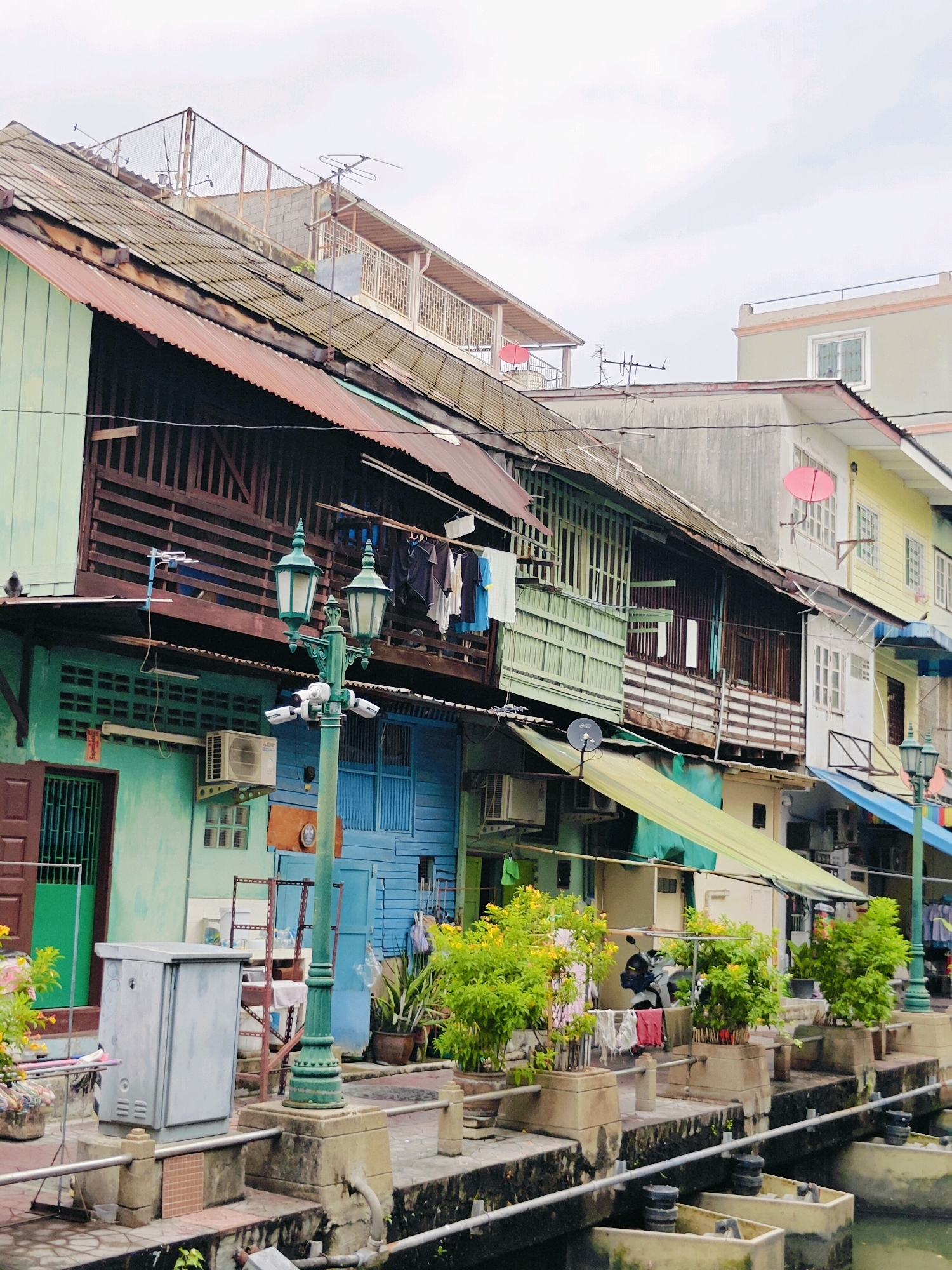 The image depicts a weathered, multi-story structure located along a canal in Bangkok, Thailand. The building exhibits a mix of architectural styles and materials, with corrugated metal roofs and wooden balconies, painted in various colors including green, blue, and yellow. Laundry hangs from a balcony on the upper floor. A street lamp with multiple lights stands in the foreground, equipped with security cameras. In front of the building, plants in pots line a walkway beside the canal, and a gray utility box is visible. The canal itself contains concrete structures. The sky is overcast.