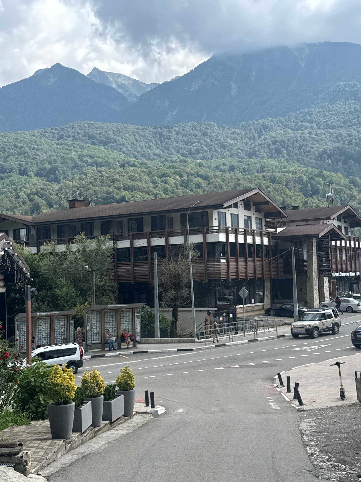 The image depicts a view of a street in Krasnaya Polyana, Russia, with the majestic Caucasus Mountains as a backdrop. In the foreground, a paved road curves, with a sidewalk on the left lined with planters containing yellow and green foliage. Across the street, modern buildings with dark wood accents and white balconies are visible, likely hotels or residential complexes. Several cars are parked or driving on the road. Bus stops with glass enclosures are situated along the sidewalk in front of the buildings. People can be seen sitting at the bus stops and walking near the buildings. The mountains in the background are covered in dense green forest at the base, transitioning to rocky peaks with a cloudy sky overhead. The overall scene suggests a resort town environment.