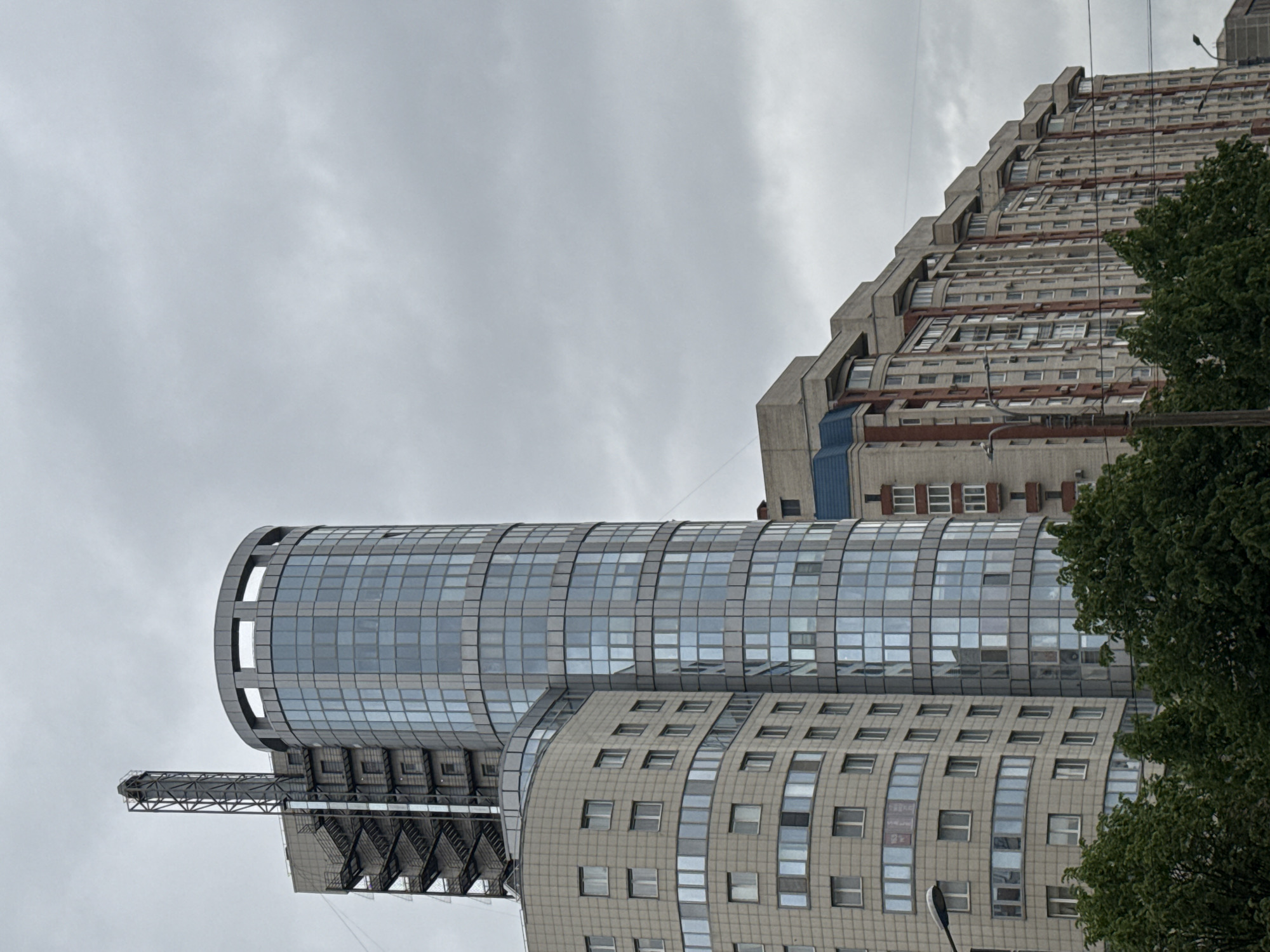 The image shows a view of several buildings in Saint Petersburg, Russia, under an overcast sky. In the foreground is a cylindrical glass building, possibly an office or commercial structure, attached to a lower, curving building with rows of windows. A metal structure, resembling a platform or framework, juts out from the side of the cylindrical building. To the right and slightly behind these buildings is a taller, stepped concrete building, likely a residential apartment block. A tree partially obscures this building. A street lamp is visible.