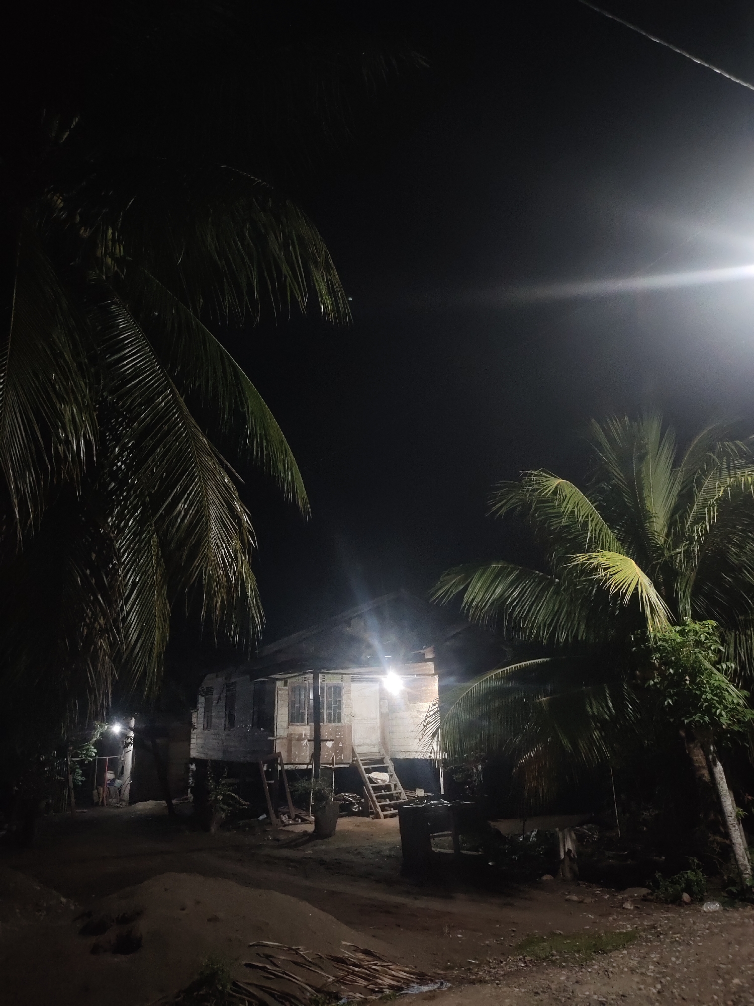 The media file captures a nighttime scene in Rayeukmatangkuli, Indonesia, dominated by a rustic, elevated stilt house constructed from wood. The house stands prominently in the center-left, with a set of wooden stairs leading up to its entrance. Multiple bright artificial lights illuminate the house, creating strong contrasts and highlights on its weathered facade, as well as casting light onto the sandy ground directly in front of it. One particularly bright light on the upper right side produces a noticeable lens flare across the dark sky.
Two large palm trees frame the scene, with one on the far left and another on the right, their fronds partially illuminated by the nearby lights. The ground is unpaved, appearing as dirt or sand, with some scattered debris like branches visible. In the background on the far left, another smaller, less distinct structure is visible, also illuminated by a bright light, and what appears to be one or two human figures can be faintly discerned in that area. The overall atmosphere is quiet and still, emphasizing the artificial illumination against the deep darkness of the night. There is no visible text in the image.