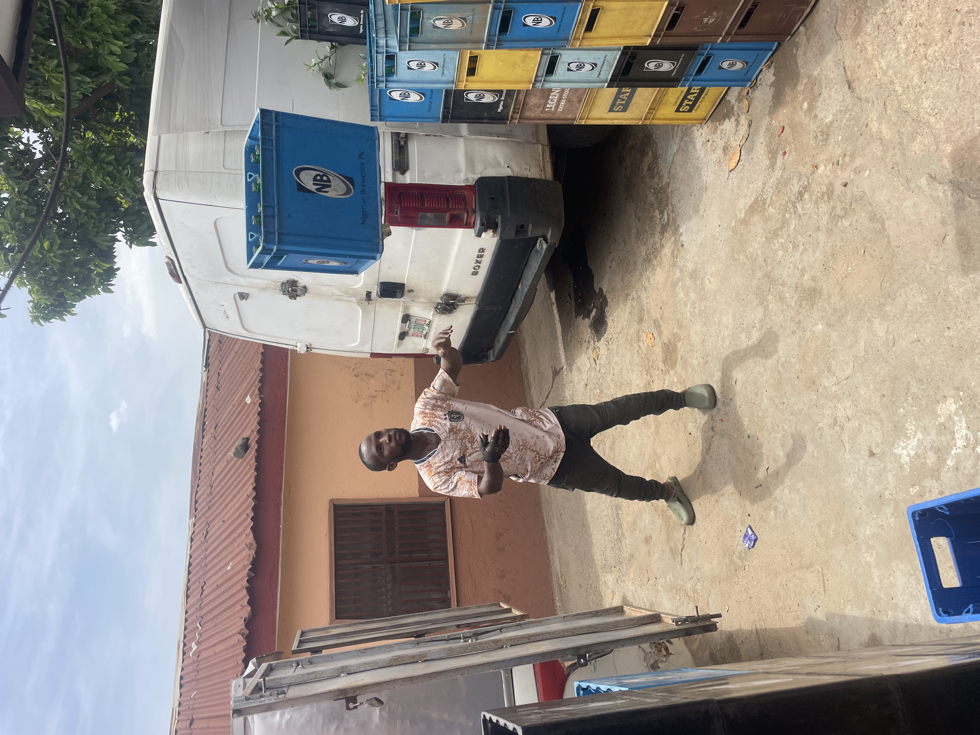 An outdoor scene in Jos, Nigeria, is depicted under bright daylight, with a man standing prominently in the foreground. He faces the viewer, wearing a light-colored, patterned short-sleeved shirt, dark trousers, and green rubber boots. His right arm is raised, and his hand is gesturing. A small, crumpled blue item lies on the light-colored concrete ground near his feet.
Behind the man, a white commercial van, identified by the word "BOXER" on its rear, is parked. The van is surrounded and possibly loaded with numerous plastic crates of various colors, including blue, yellow, black, and brown. Many of these crates prominently display the "NB" logo and the text "Nigerian Breweries Plc." Other visible brand names include "STAR," with accompanying text "Cold Filtered STAR" and "Enjoy the Brighter," and "LEGEND EXTRA STOUT." One blue crate is affixed to the back of the van.
In the background, a building with a red corrugated iron roof and peach-colored walls is visible, featuring a window with dark security bars. Green foliage from an overhead tree is also present, casting shadows that suggest a midday to early afternoon time. The ground is a rough concrete surface, showing some wear and stains. Metal frames or sheets are partially visible in the lower left foreground. The scene portrays an active distribution or storage area, likely for beverages.