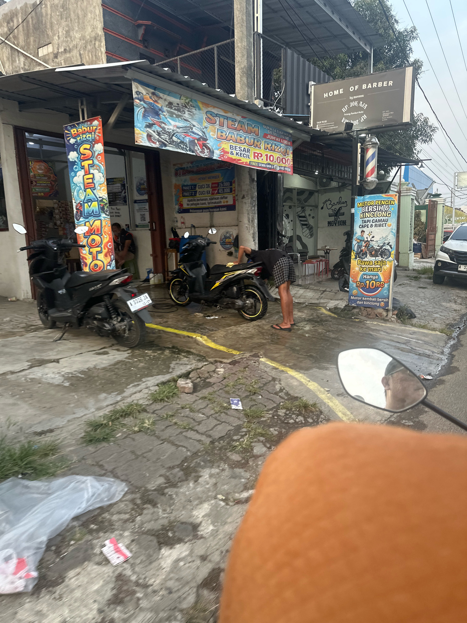 The image depicts a street scene in Saruni, Indonesia, featuring a motorcycle wash and barbershop. Two motorcycles are visible, one black and one yellow, parked on a wet, paved surface. A person wearing a black t-shirt and checkered shorts is seen washing the yellow motorcycle. A vertical banner with colorful graphics advertises "STEAM MOTOR". Another banner further down the street promotes "HOME OF BARBER" with prices. The environment appears to be an urban commercial area with buildings and signage. The overall impression is a bustling street-side service establishment.
