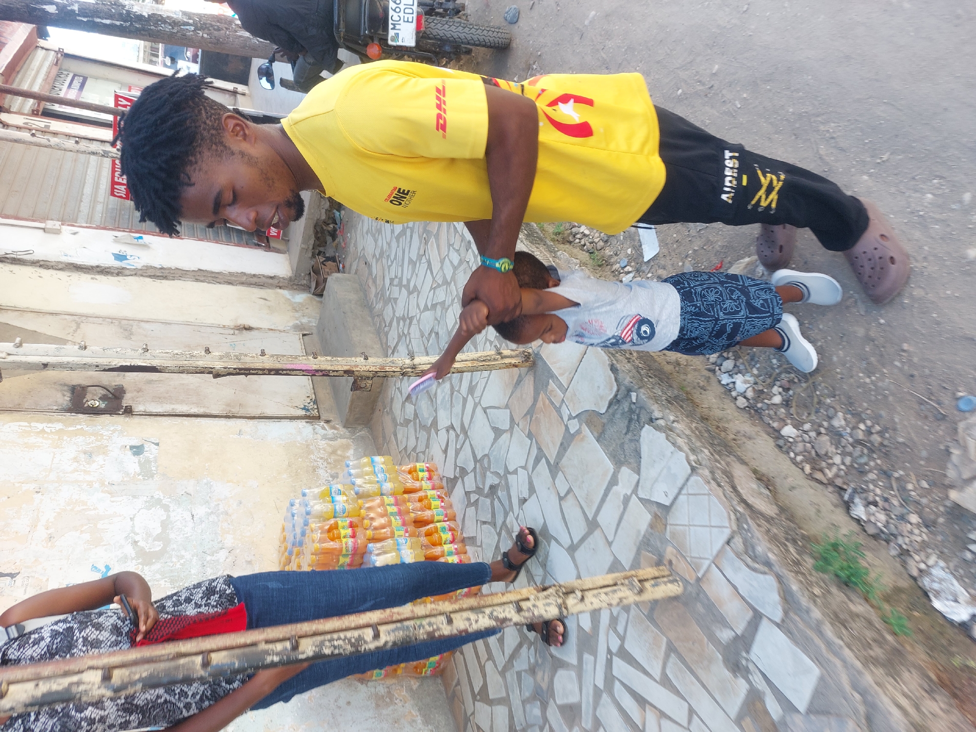 A street scene in Dar es-Salaam, Tanzania, shows a man in a yellow DHL t-shirt and black pants playfully swinging a child by the arms. The man is facing the child, who is wearing a grey t-shirt and patterned shorts. A woman, partially visible, stands near stacks of bottled drinks, holding what appears to be a mobile phone and a long, weathered piece of wood or a plank. The ground is a mix of paved tiles and dirt. In the background, there is a building with shuttered windows. A motorcycle is parked nearby.