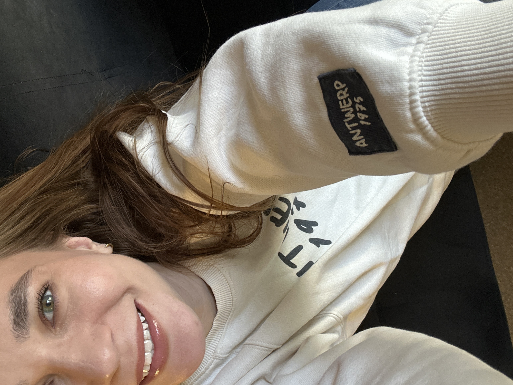 A selfie of a fair-skinned woman with light brown hair. She is smiling, showing her teeth, and her left eye is visible, appearing green. She wears a light-colored sweatshirt with a logo or writing partially visible on the front and a small rectangular tag on the sleeve that reads "ANTWERP 1975." The background shows a dark surface, possibly a couch or chair, and a small section of what appears to be carpeting in the upper right corner. The location is not discernable from the image.