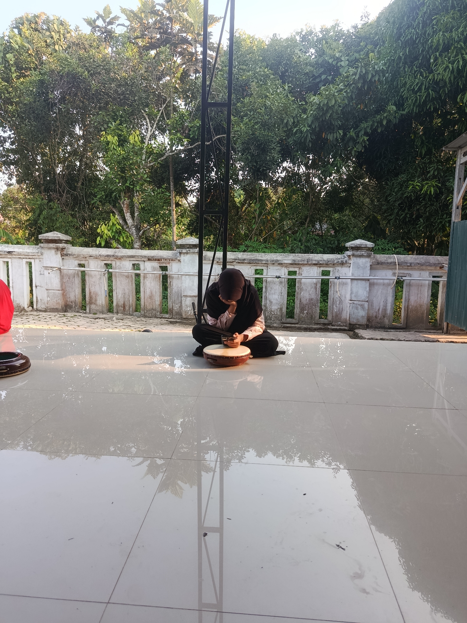 In Swarga Bara, Indonesia, a person wearing a black hijab and a light-colored top is sitting on a tiled floor, facing a wooden frame drum. The individual appears to be playing or examining the drum. The tiled floor is reflective, showing the overhead trees and a tall, dark metal structure. Behind the person, a low, white concrete wall with vertical wooden slats stretches across the scene. Lush green trees are visible beyond the wall, with sunlight filtering through their leaves. A glimpse of a red object is visible on the left side of the frame.