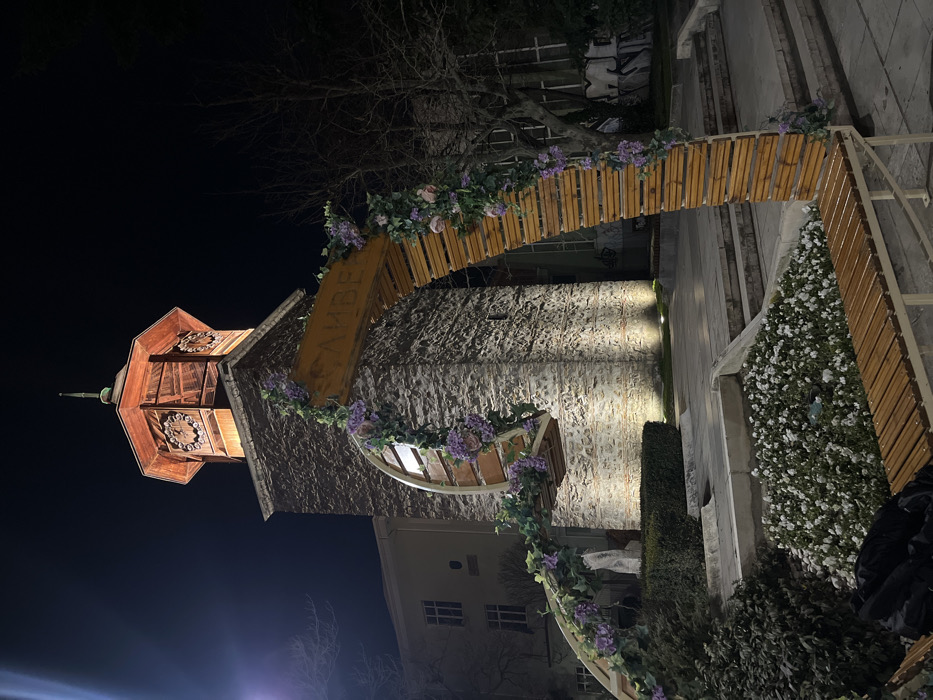 The image depicts a nighttime scene in Sliven, Bulgaria, featuring the illuminated Sliven Clock Tower. The clock tower stands centrally, constructed of rough-hewn stone at its base, which is lit by upward-facing white light fixtures. The tower's octagonal upper section, crafted from wood, contains clock faces on visible sides and is illuminated with a warm orange glow, culminating in a slender spire against the dark sky.
In the foreground, a decorative archway, constructed from light-toned wooden slats, spans from the lower right toward the left. This arch includes a curved, suspended section and is heavily adorned with artificial green foliage and clusters of purple and pink flowers. A wooden sign, bearing the Cyrillic text "СЛИВЕН" (Sliven), hangs from the archway. Directly beneath this arch, a wooden slatted bench with light-colored metal supports is positioned. Adjacent to the bench and archway, a flower bed is visible, containing numerous small white flowers. Further landscaping includes manicured green hedges and concrete steps and platforms.
In the background, on either side of the tower, dimly lit building facades are visible. Bare-branched deciduous trees stand against the dark, clear night sky. No individuals are present in the scene.