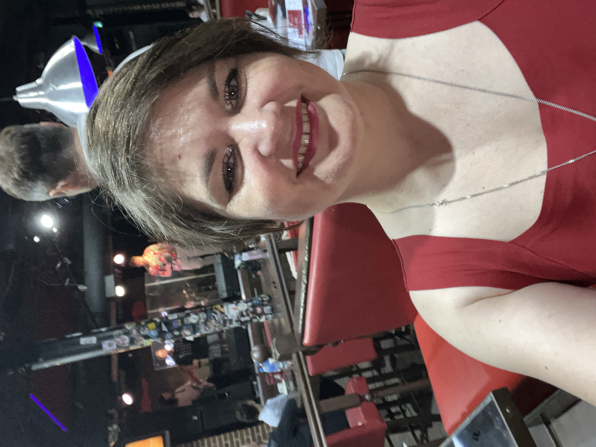 A woman in a red top smiles for a selfie in what appears to be a dimly lit restaurant or bar with a stage in the background where a band is performing. Several people are visible in the background, seated at tables or standing near the stage. The woman has short brown hair and is wearing a silver necklace. Red chairs are visible in the foreground and middle ground. The overall impression is of a casual evening outing.