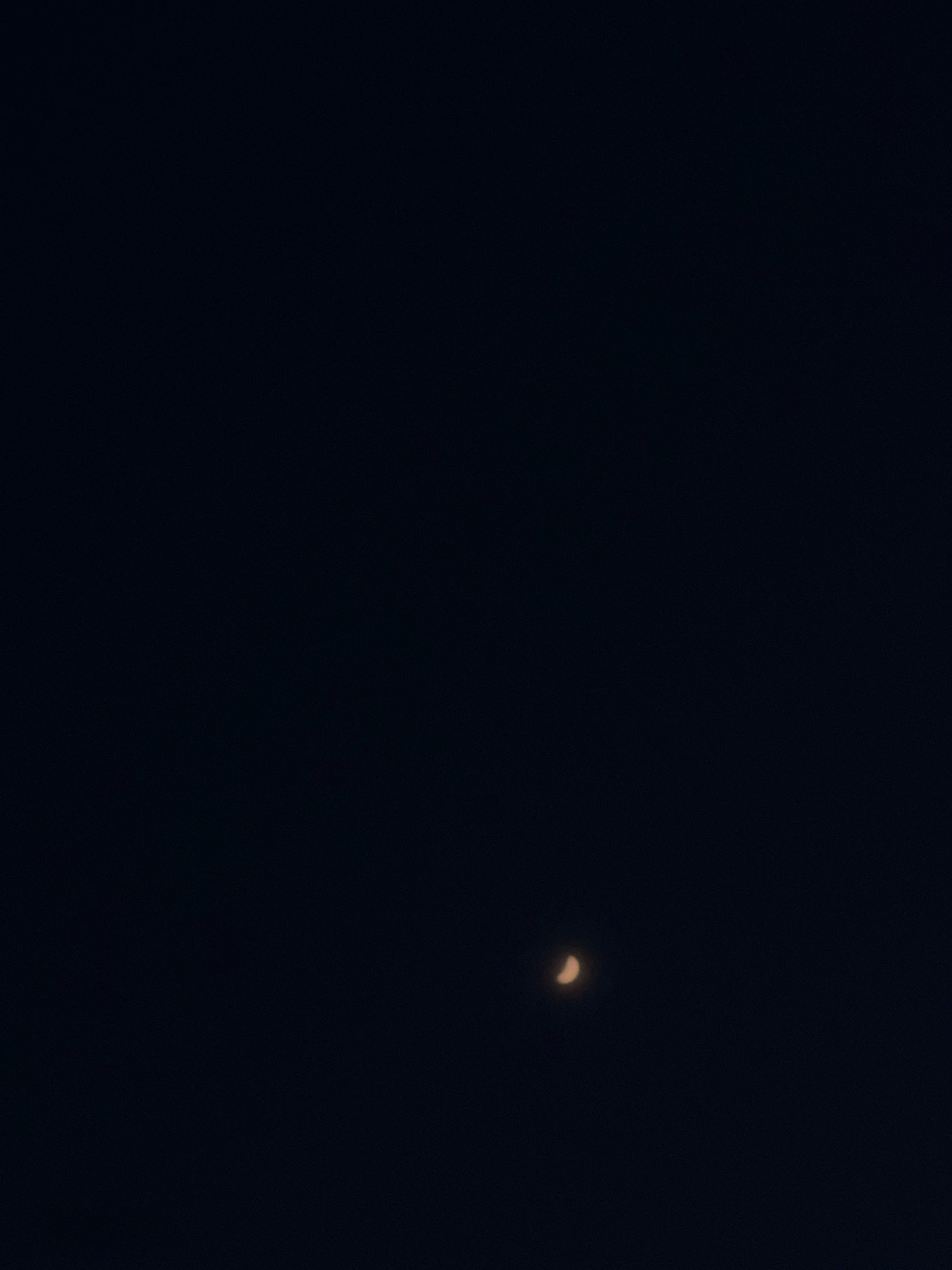 A crescent moon is visible in the dark night sky. The moon is illuminated on the right side and appears to be a pale orange or yellow color. The background is a deep, uniform black, suggesting a clear night with no other celestial bodies or light pollution readily apparent.
