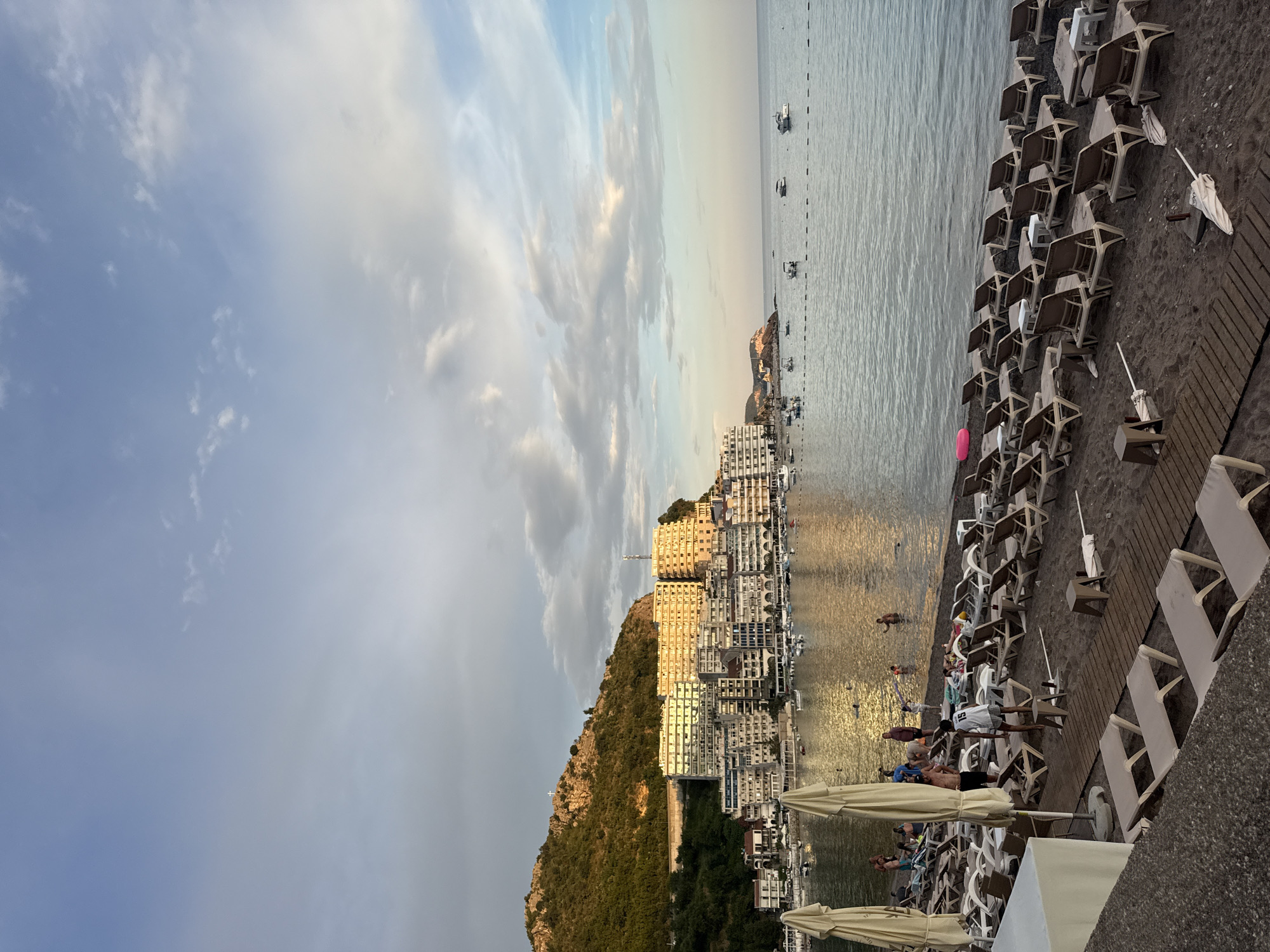 The image depicts a scenic view of a beach and a coastal town, likely taken at Rafailovići, Montenegro. The scene is oriented vertically, so it appears as a landscape with the sky at the top and the ground at the bottom.
In the foreground, there is a sandy beach lined with rows of empty lounge chairs and a few scattered beach umbrellas, suggesting either early morning or late evening. Some people are visible on the beach, some relaxing on the chairs and others walking near the water. The sea is calm and reflects the soft, golden light of either sunrise or sunset. A wooden boardwalk runs along the edge of the beach.
Beyond the beach, a densely built coastal town is nestled against a steep, green, tree-covered hill. The buildings are multi-story and appear to be hotels or apartment complexes, many with balconies. A cross is visible on the peak of the hill overlooking the town and sea. Several boats are anchored in the water near the shore.
The sky is partly cloudy, with soft hues of blue, white, and hints of pink and orange, indicative of the golden hour. The overall atmosphere is peaceful and serene, suggesting a relaxing vacation setting. No visible text is present in the image.