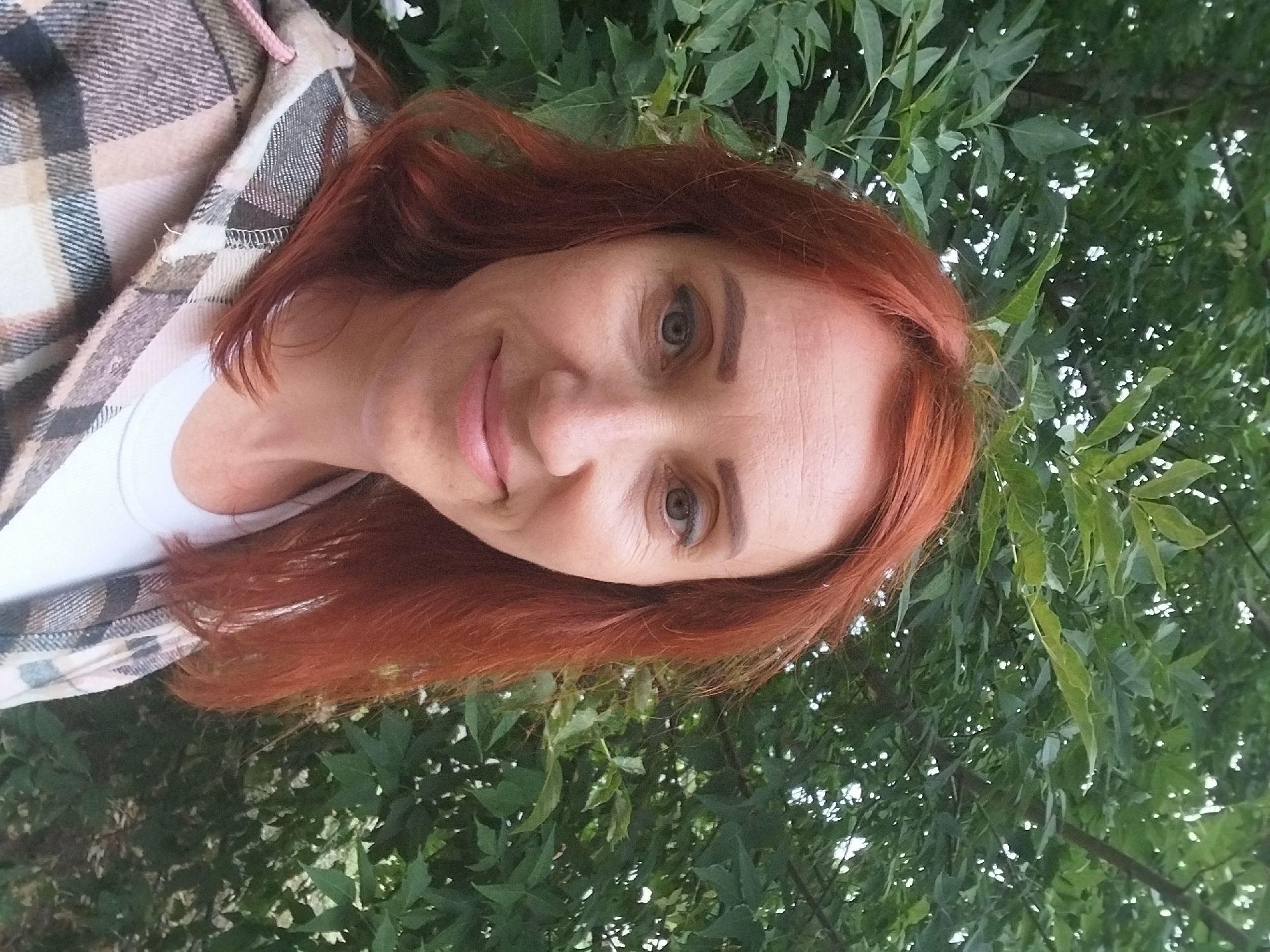 A woman with red hair is posing for a selfie outdoors in front of lush green foliage. She is wearing a white t-shirt and a plaid jacket. The image appears to have been taken in the city of Dnipro, Ukraine, based on the provided context.