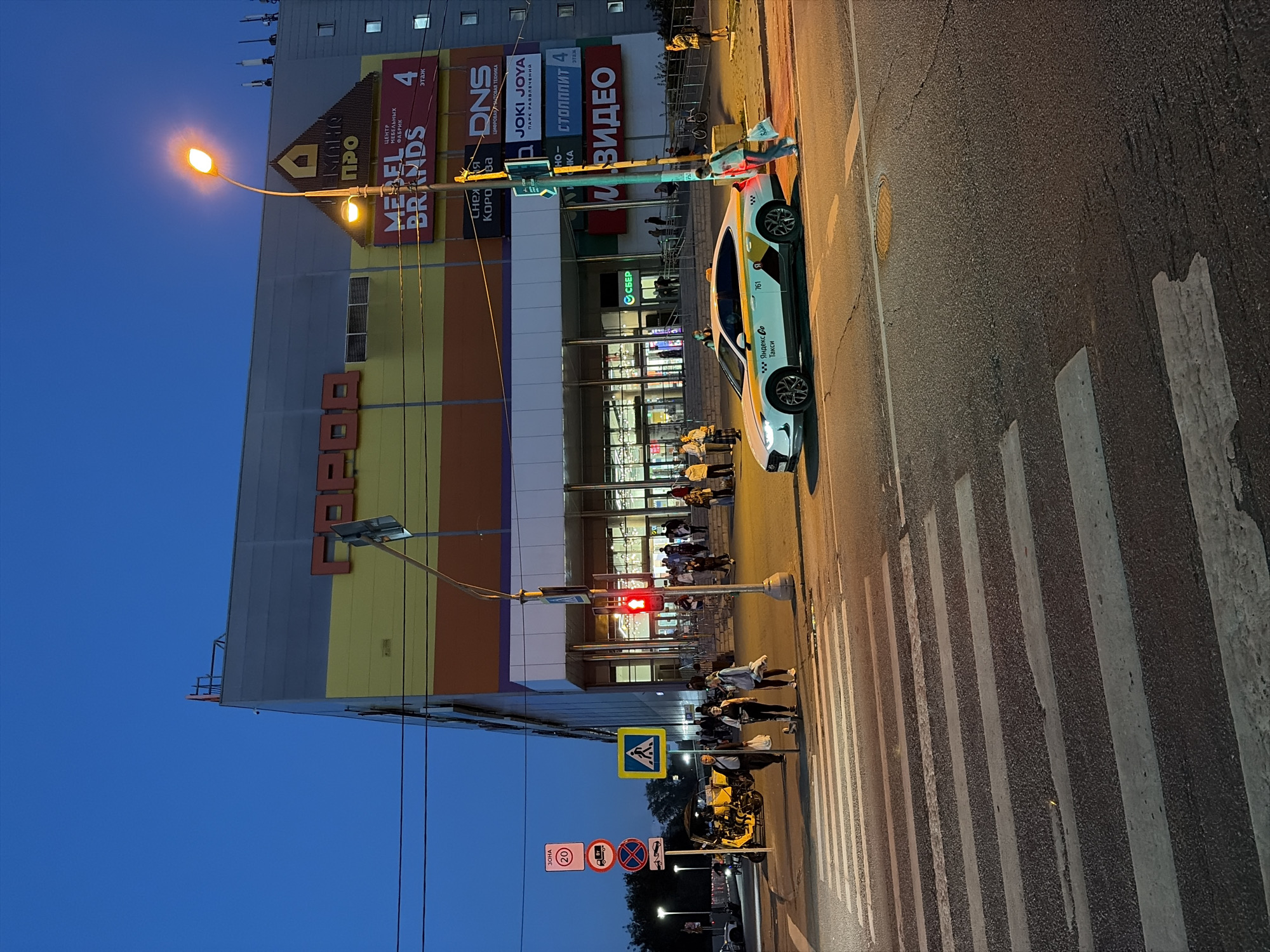 The image depicts a street scene in Moscow, Russia, at dusk. A large, multi-story shopping mall, identified by the large red lettering "ГОРОД" (Gorod) on its facade, dominates the background. The mall has multiple levels with various advertisements displayed. Notable signs include "MEBEL BRANDS", "DNS", "JOKI JOYA", "СТОПЛИТ", and "ВИДЕО". The mall has a wide glass entrance with people entering and exiting.
In the foreground, a pedestrian crosswalk is visible. A white and yellow taxi with the branding "Яндекс Go Такси" is stopped at the crosswalk. Several pedestrians are present. Some are walking, while others are on scooters. One person is walking away from the taxi, carrying shopping bags. A yellow scooter is parked on the side of the road near some traffic signs.
Traffic lights are visible, with one showing red. The sky is a deep blue, indicating the transition from day to night. Streetlights are illuminated, casting a warm glow on the scene. The overall environment is that of a busy urban area at the end of the day.