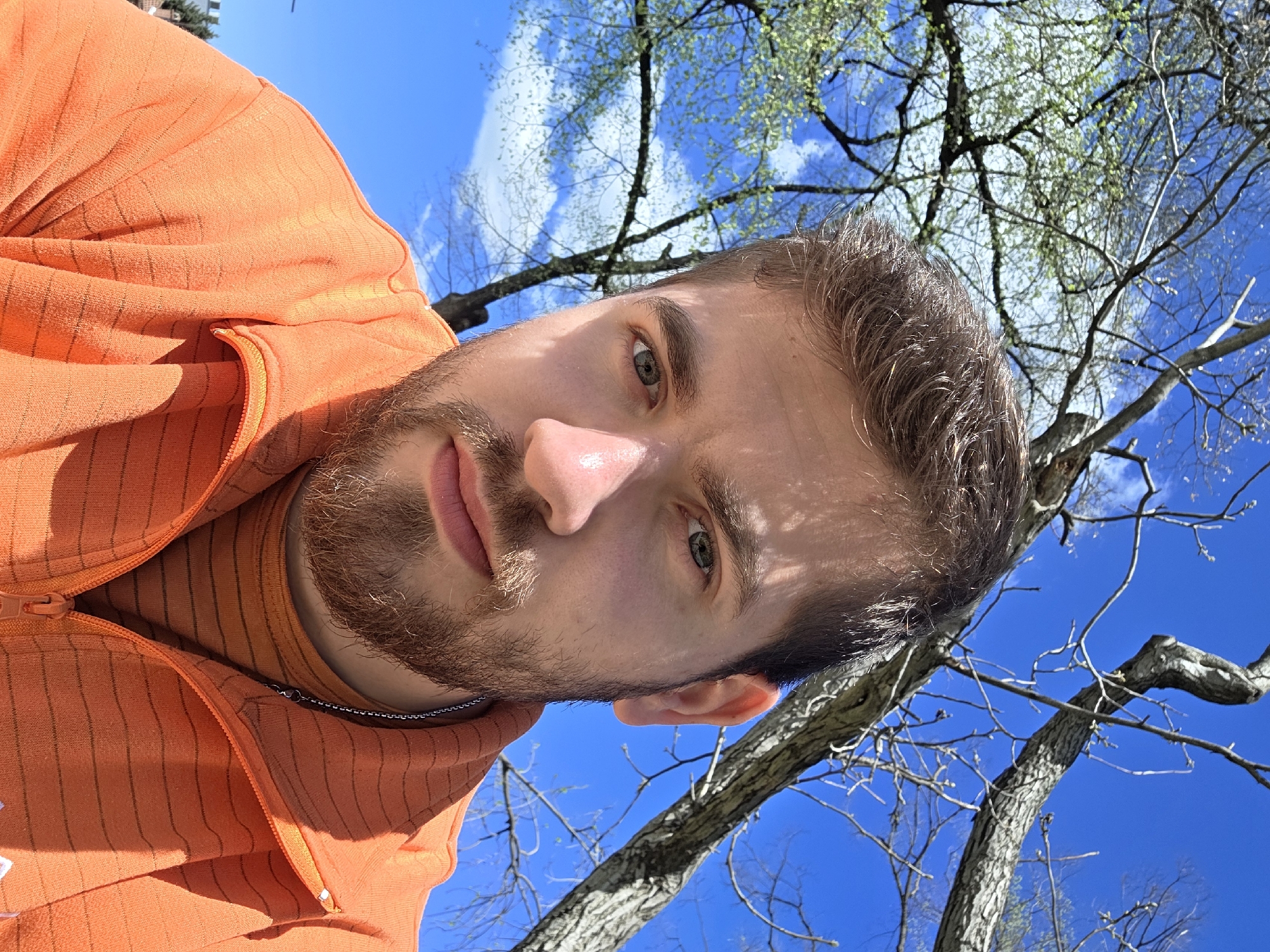 A close-up, slightly rotated selfie of a man with a light brown beard and hair, wearing an orange hooded jacket and a matching T-shirt underneath. He is facing the camera with a serious expression. The background consists of a clear blue sky with some white clouds, interspersed with branches of trees with budding leaves. The location is likely outdoors, possibly in or near a park or garden within the city of Plovdiv, Bulgaria, given the presence of trees and sky. The man is holding the camera above and to the side of his head.