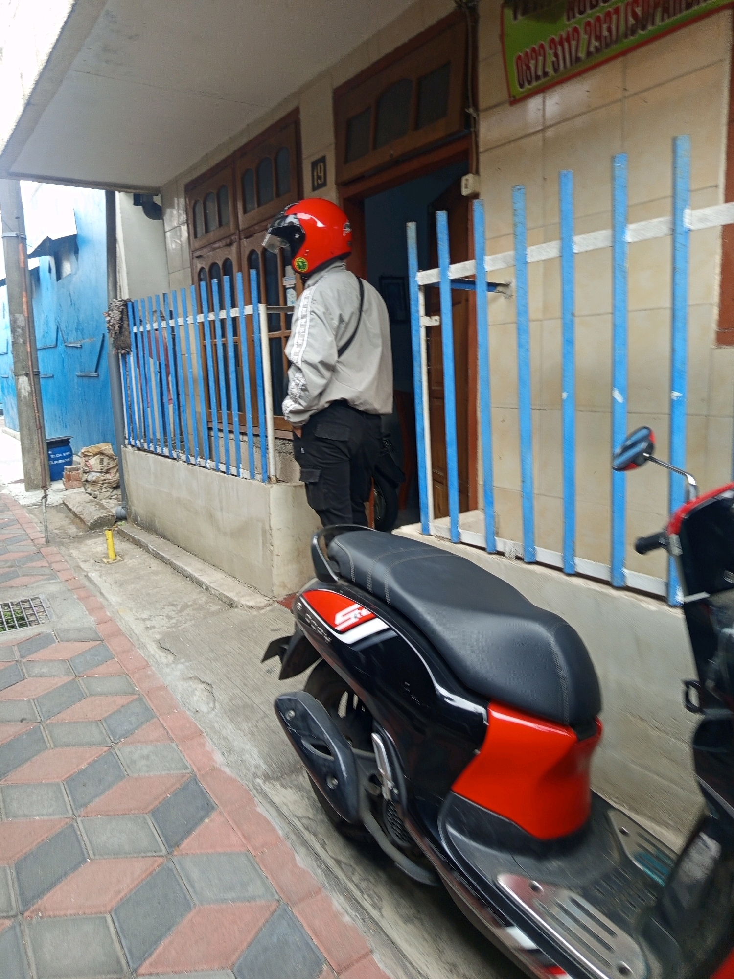 A man wearing a red helmet and a light-colored jacket stands in front of a building with a blue fence. The man appears to be facing away from the camera. A black and red scooter is parked in the foreground, partially obscuring the man and the building. The scene takes place on a paved walkway with a patterned brick design. The building has a wooden door and tiled walls. A sign with Indonesian text is visible above the door. The environment appears to be an urban setting, potentially in the city of Malang, Indonesia, as indicated by the context.
