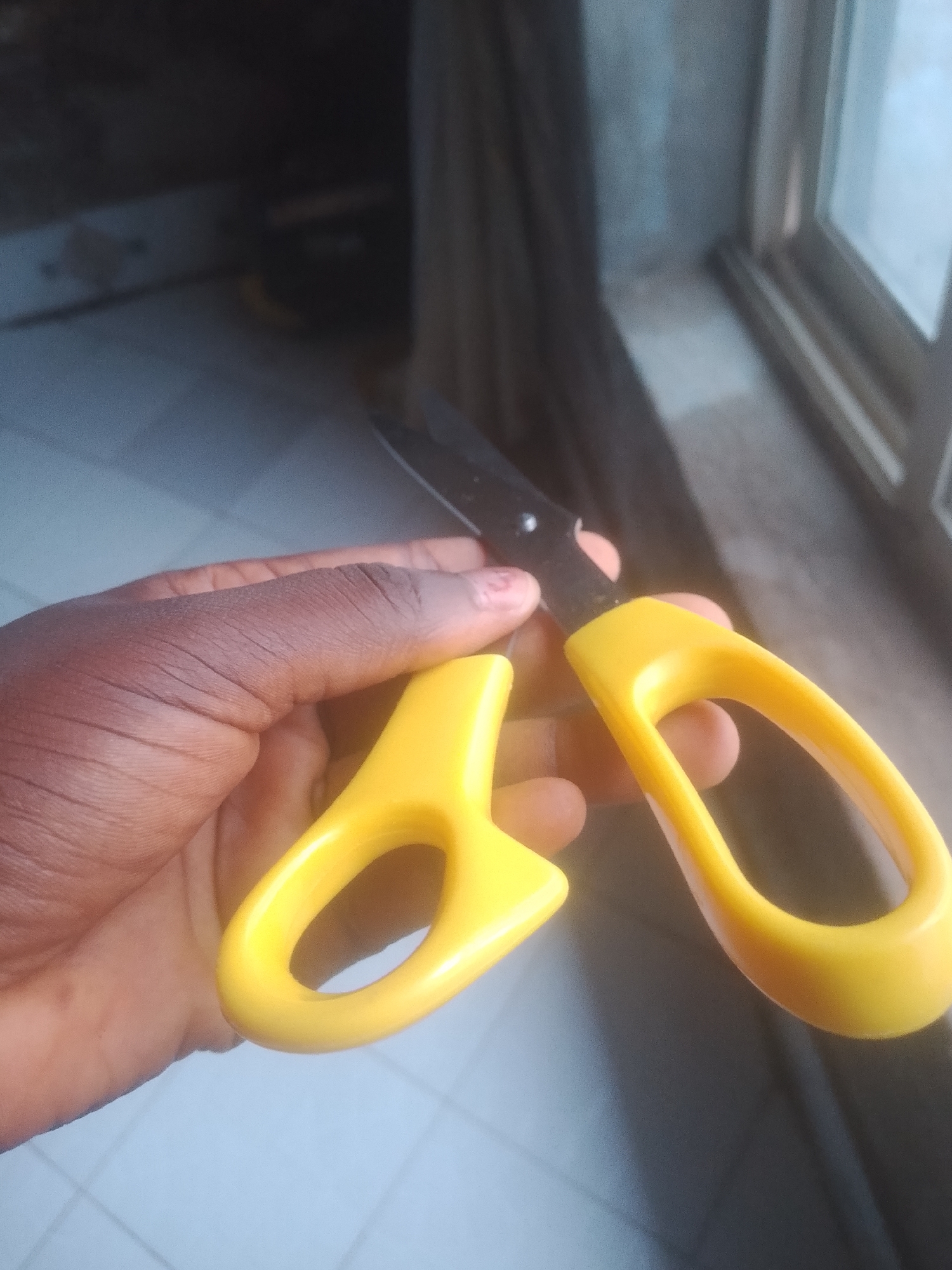 A close-up shot shows a person's hand holding a pair of yellow-handled scissors. The scissors have black blades, and the hand appears to be a dark-skinned hand. The background is blurred but suggests an indoor environment with a window. The image is taken in Jauro Yinu, Nigeria.