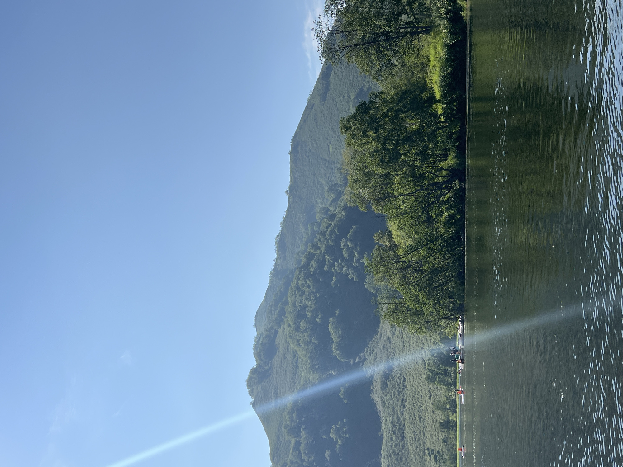 A serene lake scene in Paratunka, Russia, unfolds under a clear blue sky. The water's surface, dark and reflective, mirrors the lush green trees lining the shore and the majestic, tree-covered mountains in the distance. A prominent sunbeam cuts diagonally across the image, adding a ethereal glow. On the lake, several individuals are engaged in various water activities. Some are on paddleboards, while others are gathered on a floating dock, suggesting a leisurely summer day. The overall impression is one of tranquility and natural beauty.