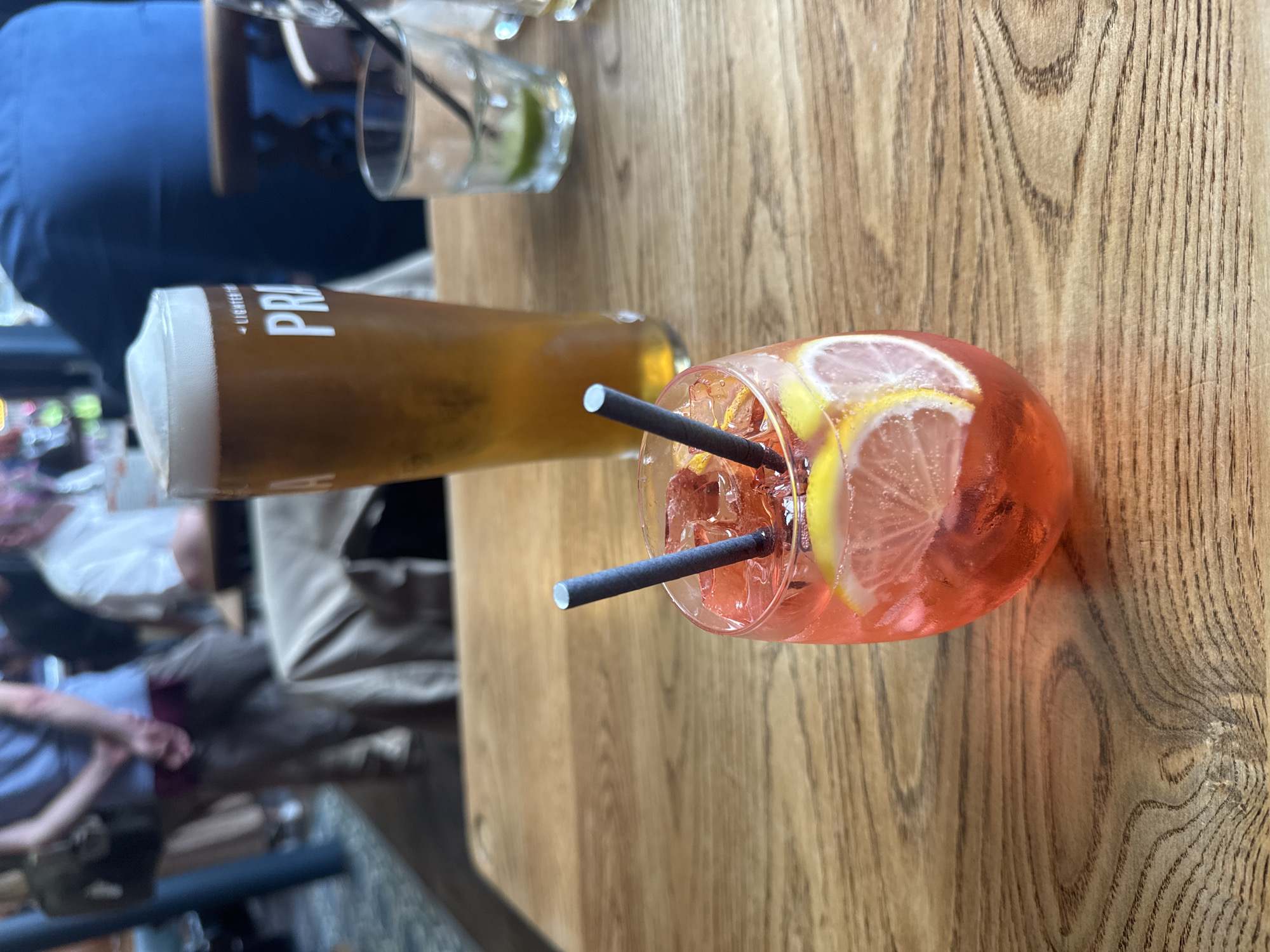 This is a medium-shot, eye-level, indoor photograph of two drinks on a wooden table, likely at a bar or restaurant. In the foreground, a pink cocktail in a round glass is filled with ice, lemon slices, and two dark straws. In the background, a tall glass of beer with a frothy head sits to the left, and a smaller glass with ice and a lime slice is visible to the right. Out of focus figures of people are in the background, suggesting a social setting. The wooden table has a prominent grain pattern. The overall lighting is warm and ambient. The photograph does not contain any specific landmarks or contextual information to determine the location as Glasgow, United Kingdom.
