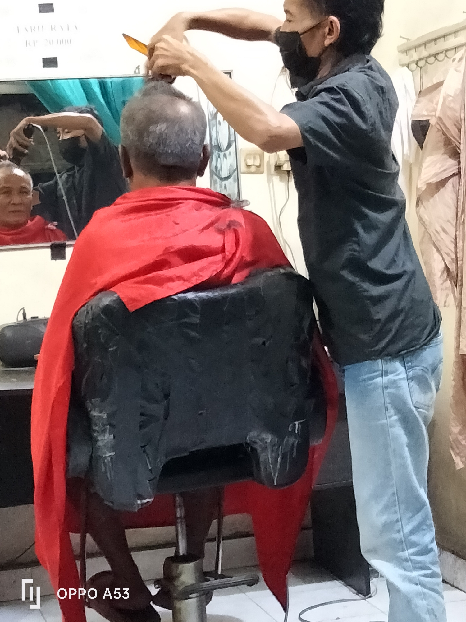 A person is having their hair cut by a barber in Denpasar, Indonesia. The person receiving the haircut is seated in a chair and draped in a red cape. The barber, wearing a black shirt and jeans, is using scissors to cut the person's hair. The scene appears to be in a barbershop, with mirrors and hair-cutting equipment visible. The overall setting is dimly lit and suggests a humble establishment.