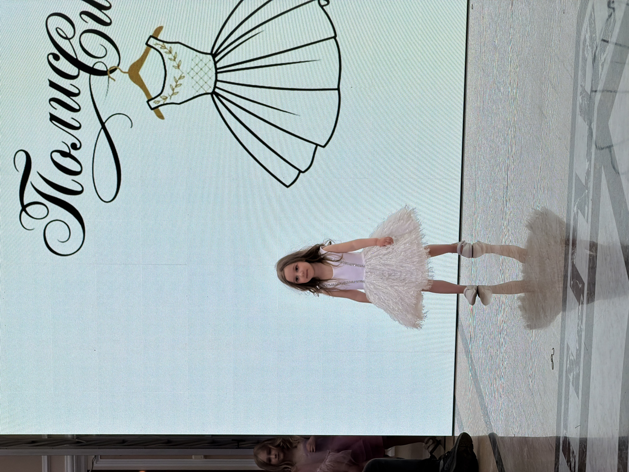 A girl in a white dress and white shoes is standing in front of a display in Saint Petersburg, Russia. The display features a logo of a dress and the text "Полиция стиля". The girl's reflection is visible on the floor. At the bottom of the image, two adults are partially visible.