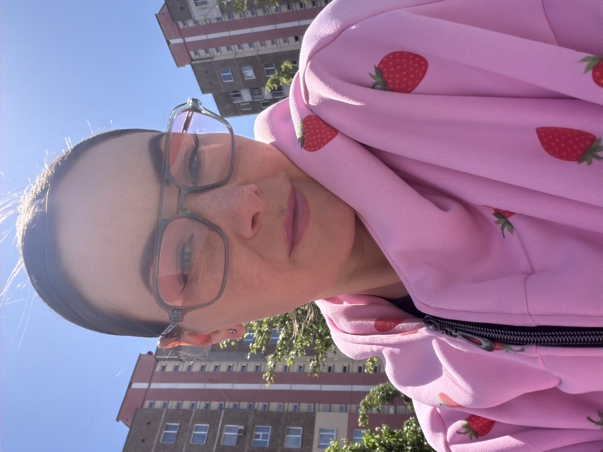 A woman with brown hair pulled back is wearing eyeglasses with clear frames and a pink zippered hoodie with a strawberry pattern. Buildings are visible in the background, along with trees. The location is a city; Unknown, Unknown.