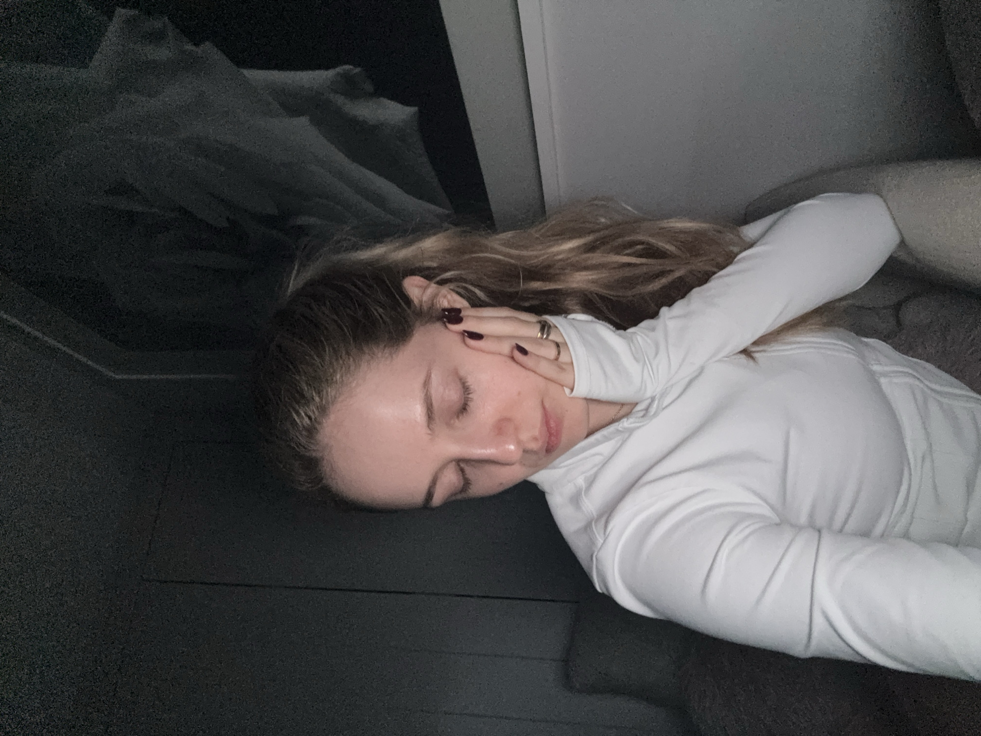 A young woman with light skin and light brown hair is lying down, eyes closed, with her left hand touching her face. She is wearing a white, long-sleeved shirt. The background appears to be dark, with a grey wall behind her and bedding to the left. The location is not discernible from the image.
