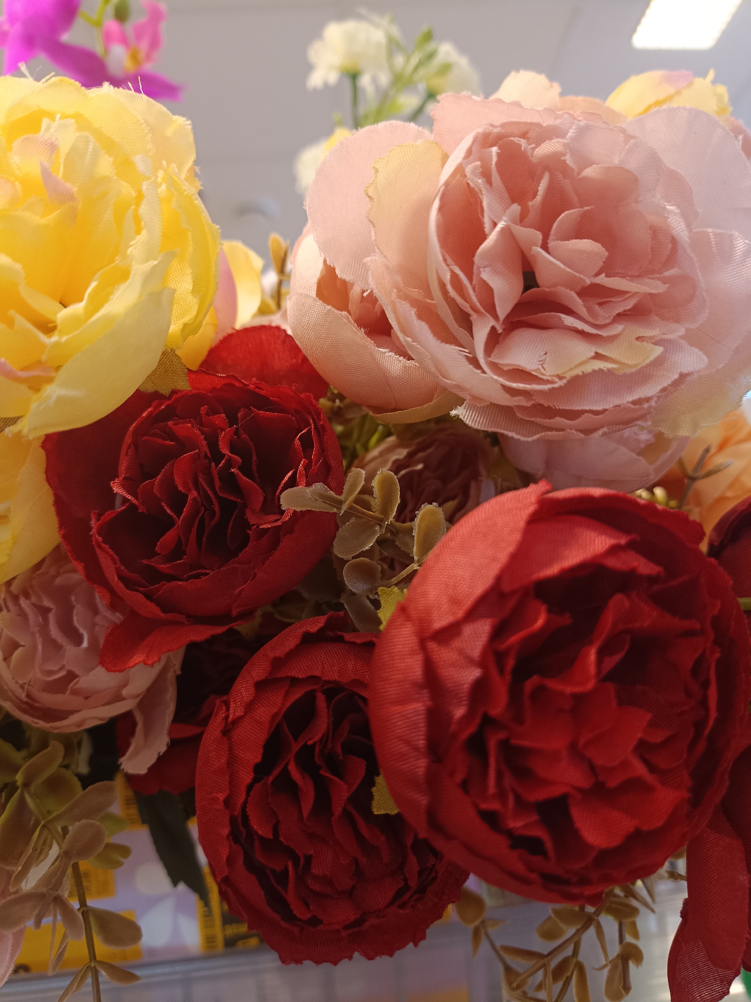 The image depicts a bouquet of artificial flowers. Dominant colors are yellow, pink, and red. The bouquet is densely packed, featuring various rose-like blossoms. Some small, green, artificial leaves are visible among the flowers. The background is slightly blurred and appears to be indoors, possibly with artificial lighting. No people or specific location markers are present in the image to confirm it is in Uinskoye, Russia. The image solely shows an arrangement of artificial flowers.