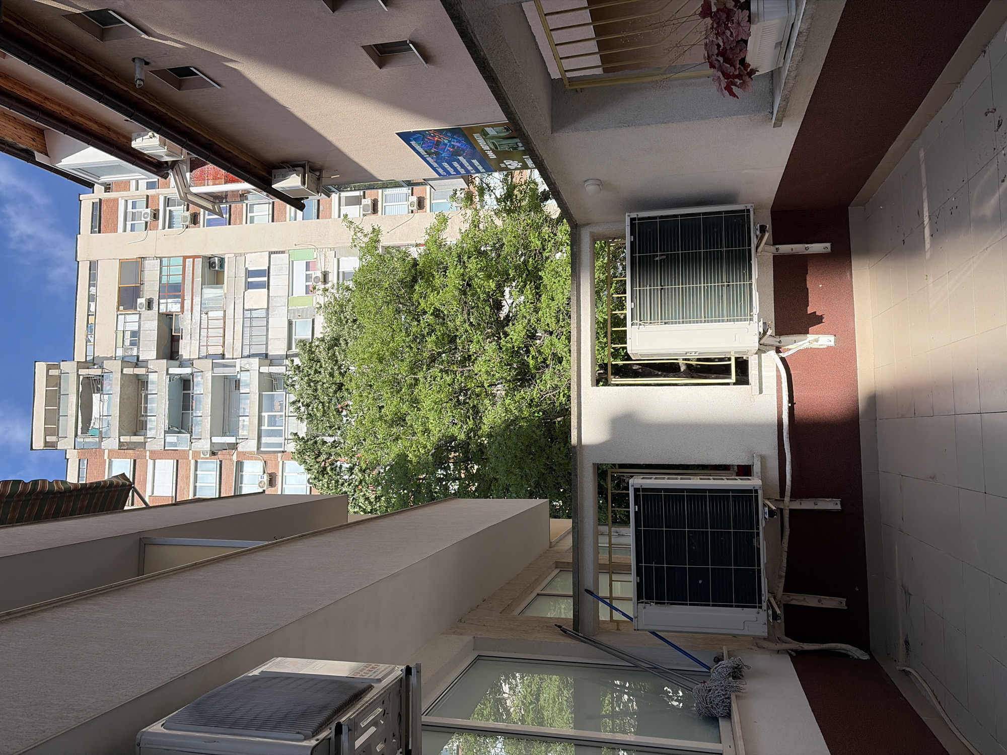The image depicts an exterior view in an urban setting, possibly in Stara Zagora, Bulgaria. The perspective is looking upwards, revealing a tall apartment building with multiple windows and balconies against a blue sky. A large, mature tree with dense foliage partially obscures the building.
The foreground shows the exterior of another building, likely part of the same complex, featuring air conditioning units mounted on the walls. One unit is visible on the right, near a red tiled wall, and another unit is on the bottom left. A window with a white frame and a partial mop head is also in the foreground.
A sign with a blue background is mounted on the ceiling connecting the buildings. The scene captures elements of urban life, with architectural features and the presence of utilities like air conditioning units.