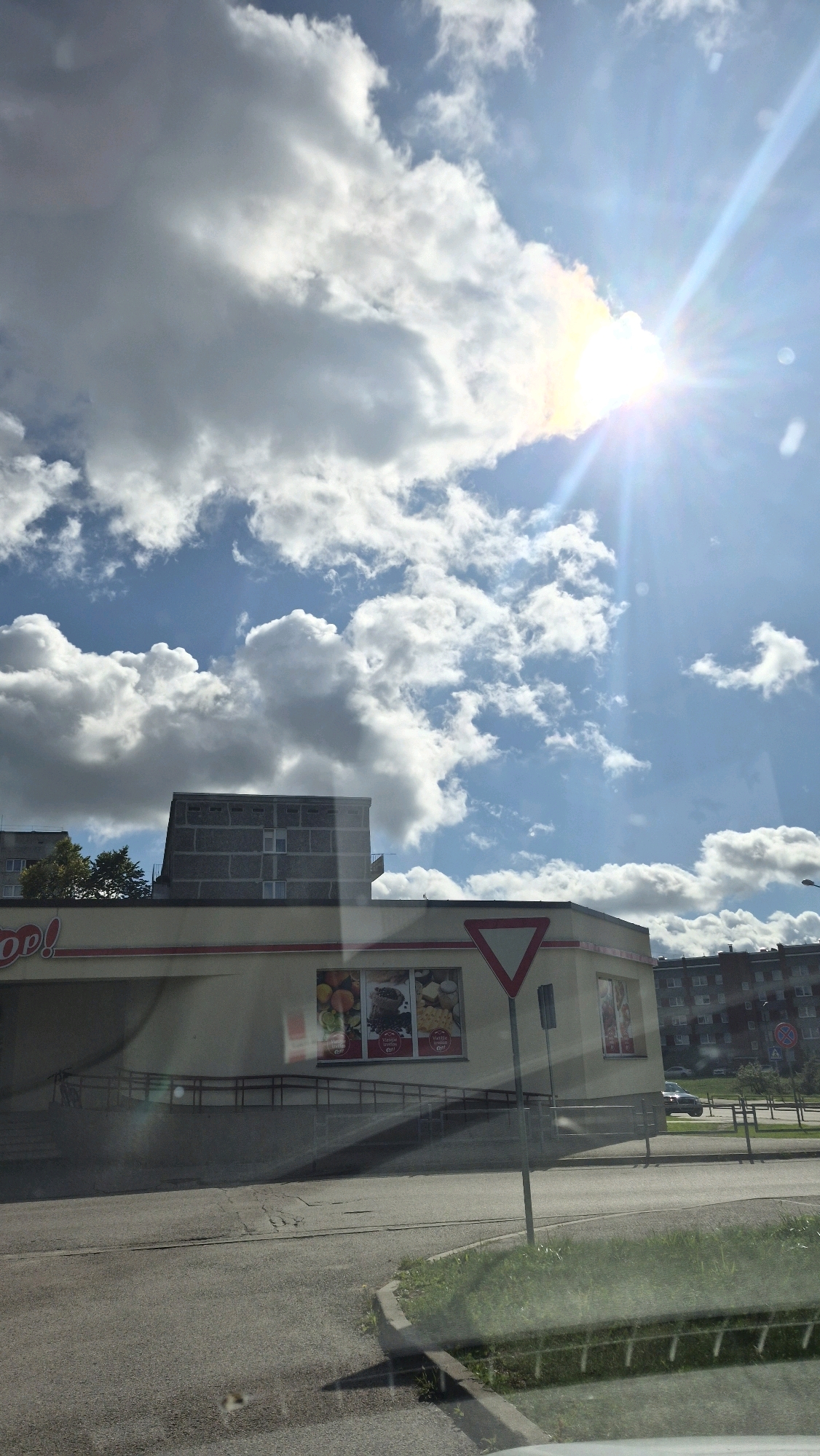 The image shows a bright, sunny day with scattered clouds. The sun is prominently visible in the upper right corner, casting a lens flare and bright rays across the sky. In the foreground, there is a street with a curb and some patchy grass. A building with a yellow facade and a red trim runs horizontally across the middle of the image. The building appears to be a store, with windows displaying advertisements for produce and other goods. A red triangular yield sign is mounted on a pole in front of the building. Behind this building, there is a tall, grey, concrete apartment building. Further in the background, more apartment buildings are visible, along with trees and streetlights. The scene appears to be an urban environment, likely in a residential or commercial area. Based on the context provided, this location is in the city of Daugavpils, Latvia.