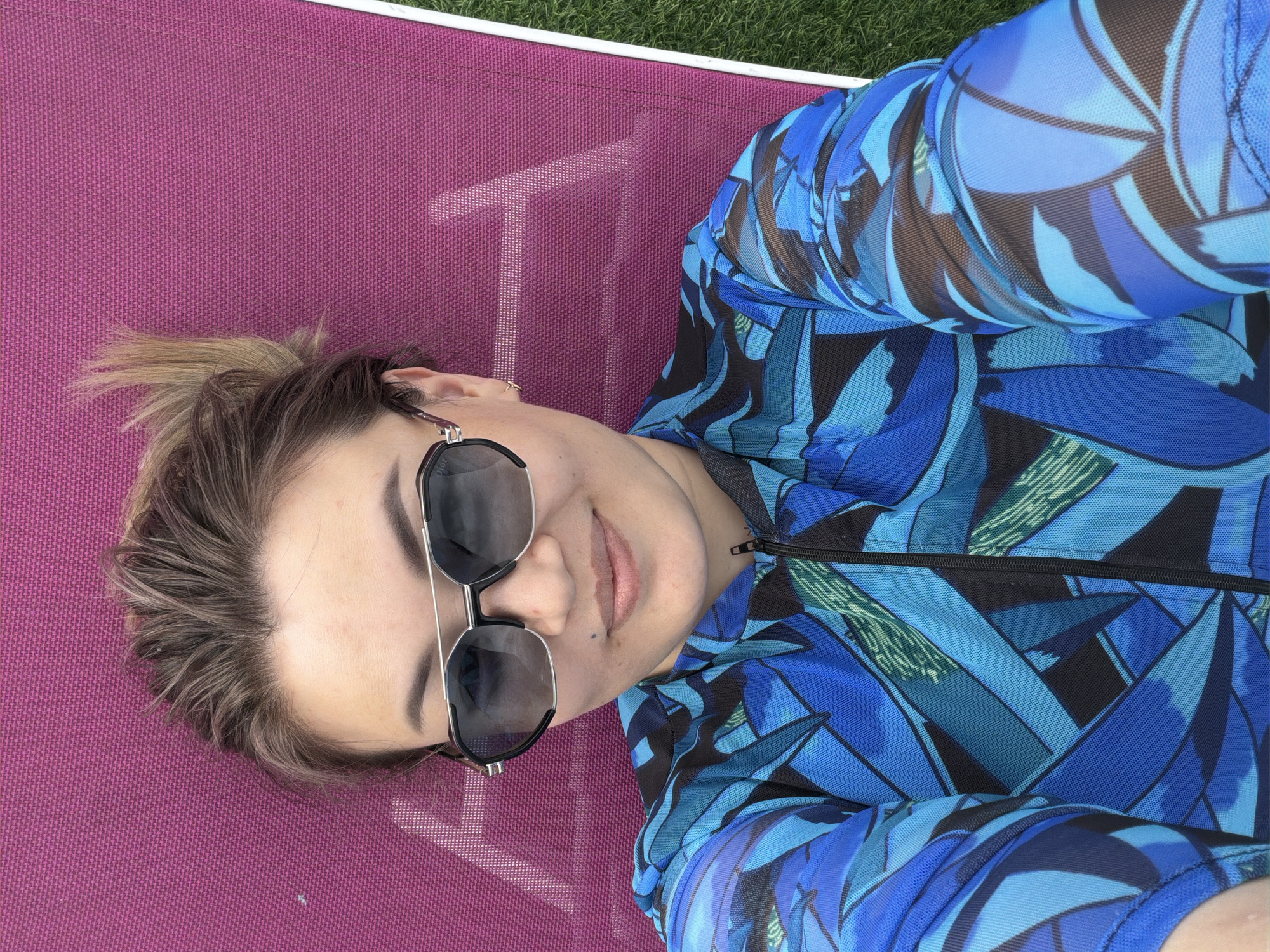 A selfie is taken from a high-angle shot of a woman lying on a magenta sun lounger. She is wearing large, dark-lensed aviator sunglasses, a blue and black patterned zip-up jacket, and has her hair tied up in a messy bun. The lounger is on a patch of green grass. The background is out of focus, but suggests an outdoor setting, possibly a park or backyard in Молбаза, Kazakhstan.