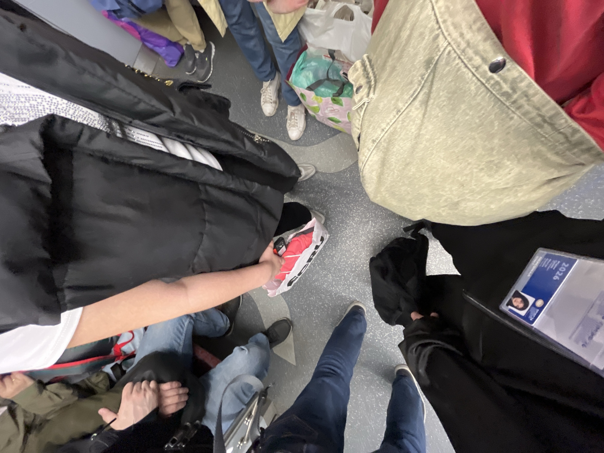 This high-angle, top-down photograph captures a group of individuals in what appears to be a crowded public transport setting, possibly in Saint Petersburg, Russia, given the provided context. The gray floor with light geometric patterns suggests an interior space like a subway car or tram.
At least five people are visible, primarily from the waist down or as disembodied limbs, wearing various casual attire including jeans in different washes (light blue, dark blue) and sneakers in white, black, and gray. One person on the left is seated, with their hands folded, wearing an olive-green jacket and blue jeans. Another person, to their right, is reaching into a white paper bag, revealing a bright red item inside.
Various bags and belongings are scattered on the floor among the individuals. A large, light green and white patterned tote bag is visible towards the top right. A black jacket or bag lies near a person's feet on the right side of the frame, next to which an ID card is prominently displayed. The ID card is blue and white, features a small photo of a person, and clearly shows the year "2026". Other faint text on the card appears to be in Cyrillic, consistent with the Russian context. The overall scene suggests everyday commuting or travel within an urban environment.