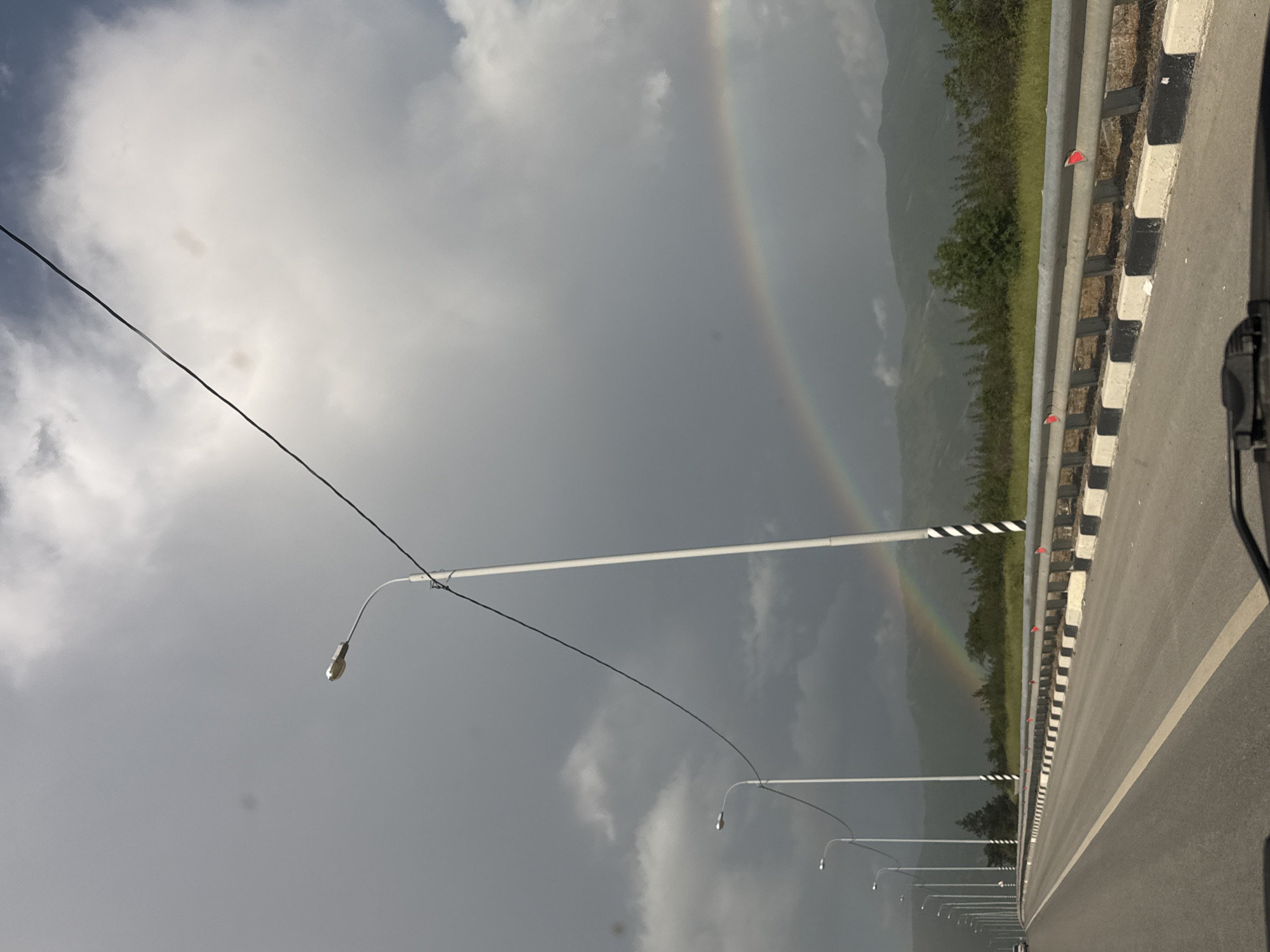 The image shows a road and a rainbow. The road is paved and has a white line running down the center. A guardrail runs along the right side of the road. There are lampposts along the left side of the road. The rainbow appears to be in the distance, against a cloudy sky. The location is near Gelendzhik, Russia.