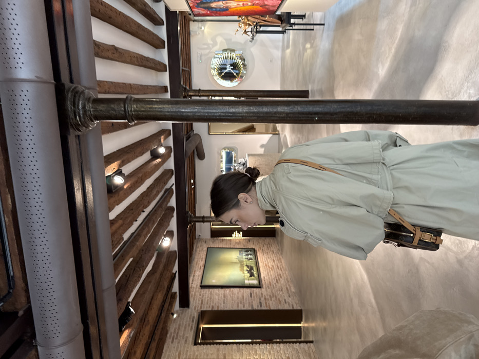 The image depicts the interior of a renovated exhibition space or art gallery, consistent with a historical building in Venice, Italy. A female subject, viewed from the rear and in left profile, stands in the mid-ground. She has dark hair tied back and wears a light beige trench coat with a dark brown patterned cross-body bag featuring tan leather straps. Her gaze is directed towards the left.
A prominent dark, fluted metal column extends from the floor to the ceiling on the right side of the frame. The ceiling above features exposed dark wooden beams set against white panels, illuminated by multiple recessed spotlights. A large, light grey perforated air duct runs horizontally across the upper ceiling. The floor is a light, smooth, uniform surface, likely polished concrete or resin.
In the left background, a brick wall displays a rectangular framed artwork depicting a tranquil waterscape with boats and distant buildings under a hazy sky. Adjacent to this, a modern rectangular doorway or recess with vertical golden-brown illuminated trim is visible. The right background features white walls adorned with two circular mirrored artworks, one exhibiting a geometric pattern. Various dark display stands hold sculptural objects, and a vibrant, partially visible portrait painting is positioned on the far right. A light-colored counter or display unit is also present in the right mid-ground.