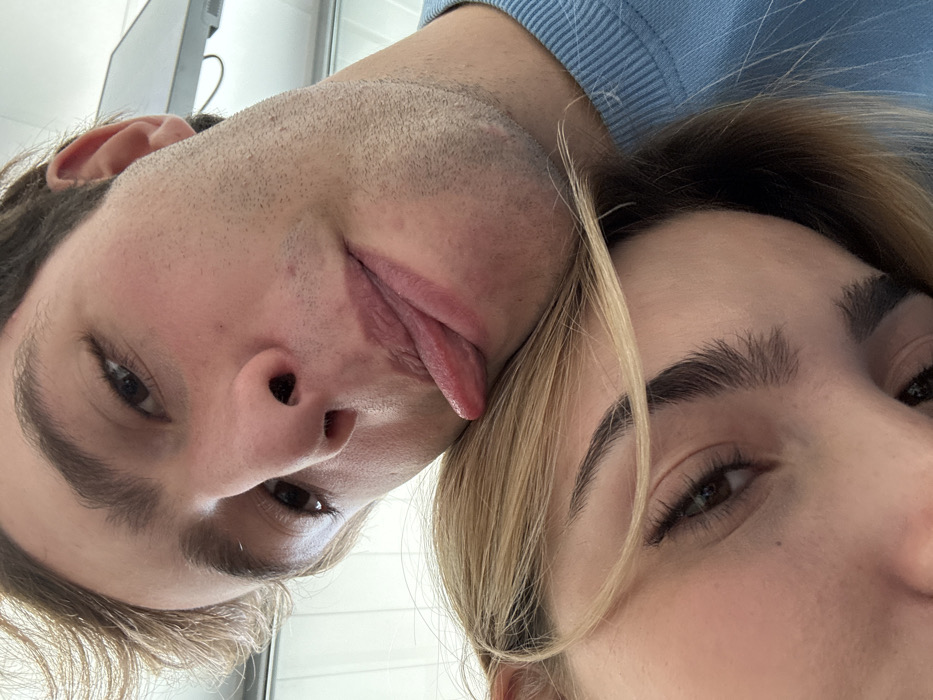 This close-up selfie depicts two individuals in Saint-Priest, France. The upper subject is a light-skinned male with short, light brown hair and visible stubble on his cheeks and chin. He wears a light blue crew-neck sweater and is seen looking towards the camera with his tongue extended from his mouth, resting his head on the lower subject's head. His right eye, brown in color, is visible. The lower subject is a light-skinned female with blonde hair that partially covers her face. Her right eye, brown with prominent eyelashes, and a well-defined eyebrow are visible. The background is uniformly light-colored and out of focus, suggesting an indoor setting, with a blurred rectangular object in the upper right corner.