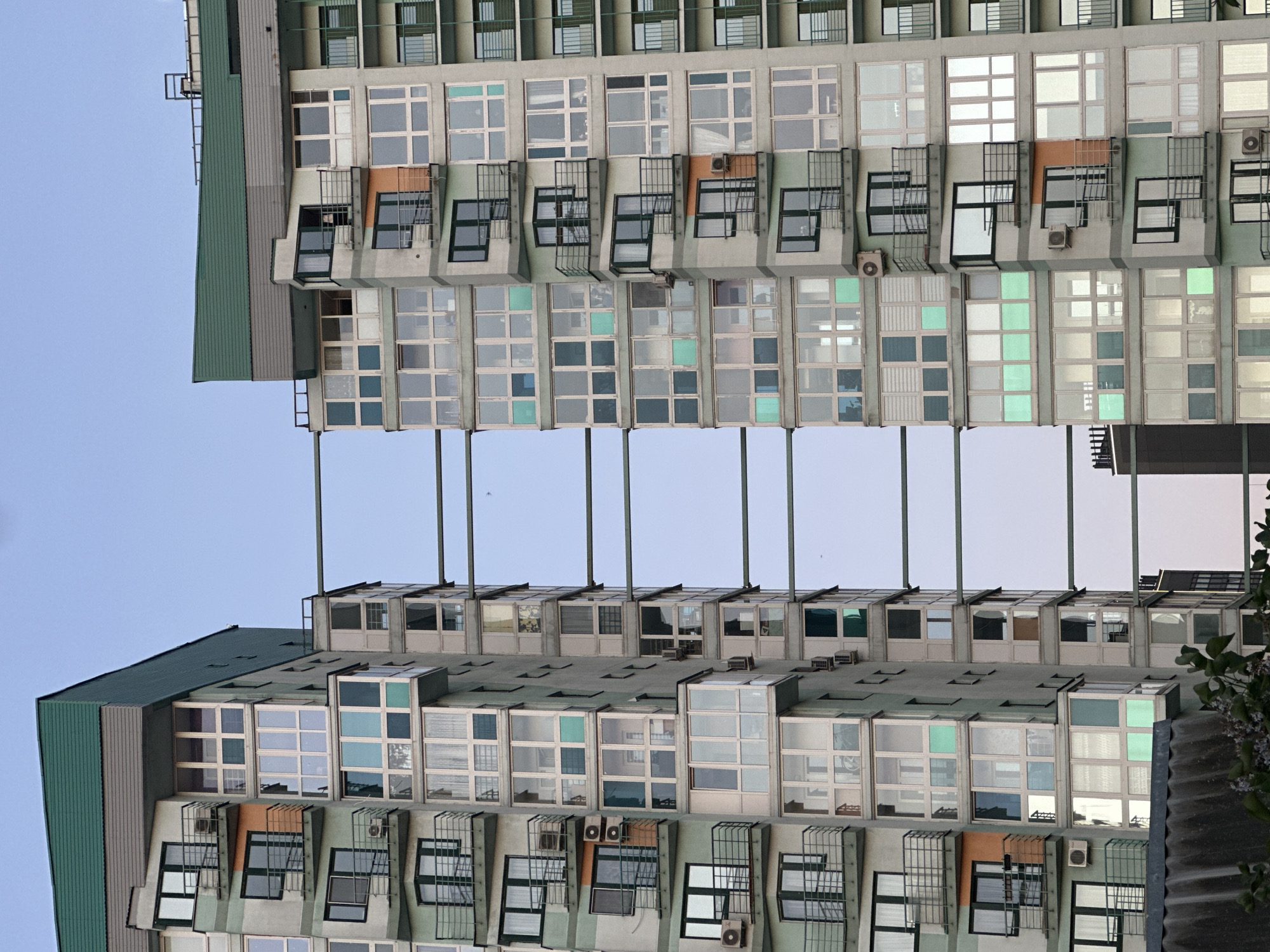 The image depicts a multi-story building in Minsk, Belarus, notable for its modernist architecture and repetitive window patterns. The building appears to be divided into modular units, each featuring multiple windows, some with open metal grates or balconies. Air conditioning units are visible beneath some windows. The building is photographed from a low angle, with the upper portion mirroring the lower part, creating a symmetrical, abstract composition. The sky is visible in the gap between the two mirrored sections. The building’s exterior features gray and green elements. The photograph's composition emphasizes the geometric forms and patterns within the building's design.