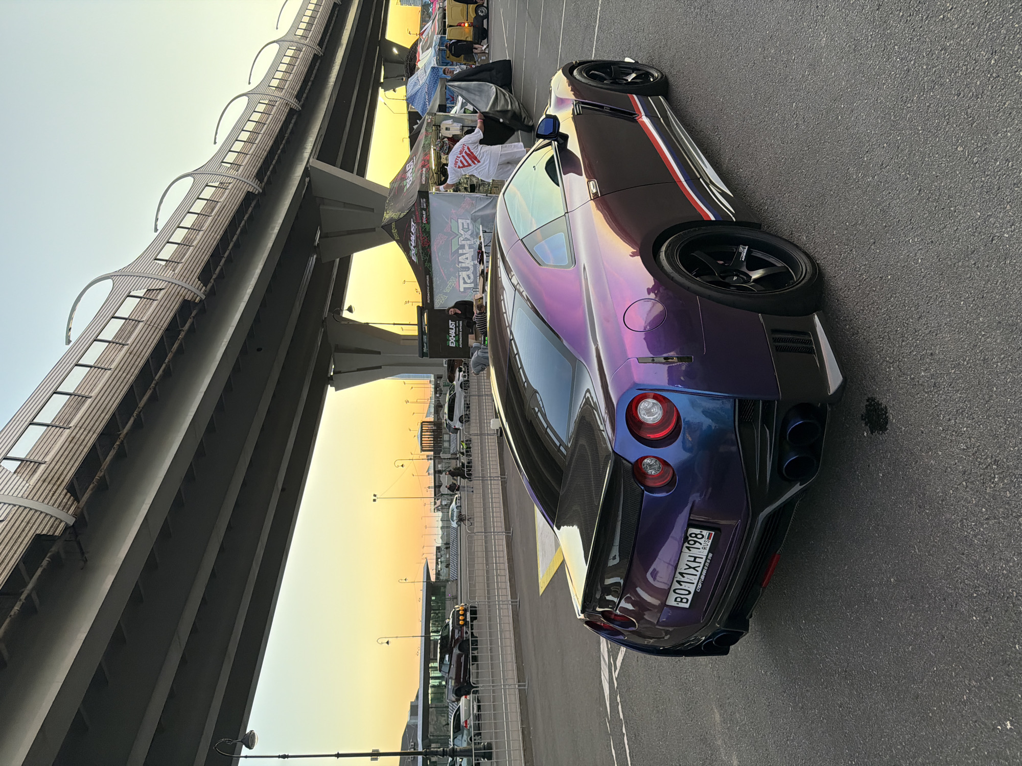 In an outdoor urban setting, a modified Nissan GT-R, painted in a shimmering purple and blue iridescent finish with red and white racing stripes along the side, is parked on asphalt. The car's rear is prominently featured, showcasing its taillights, quad exhaust pipes, and a carbon fiber spoiler. In the background, under a large overpass, several tents are set up, with some people visible interacting with them, including one person holding a large black tarp. Other vehicles, including a pickup truck and a van, are also present in the background. The sky displays a warm sunset gradient of yellow and orange.