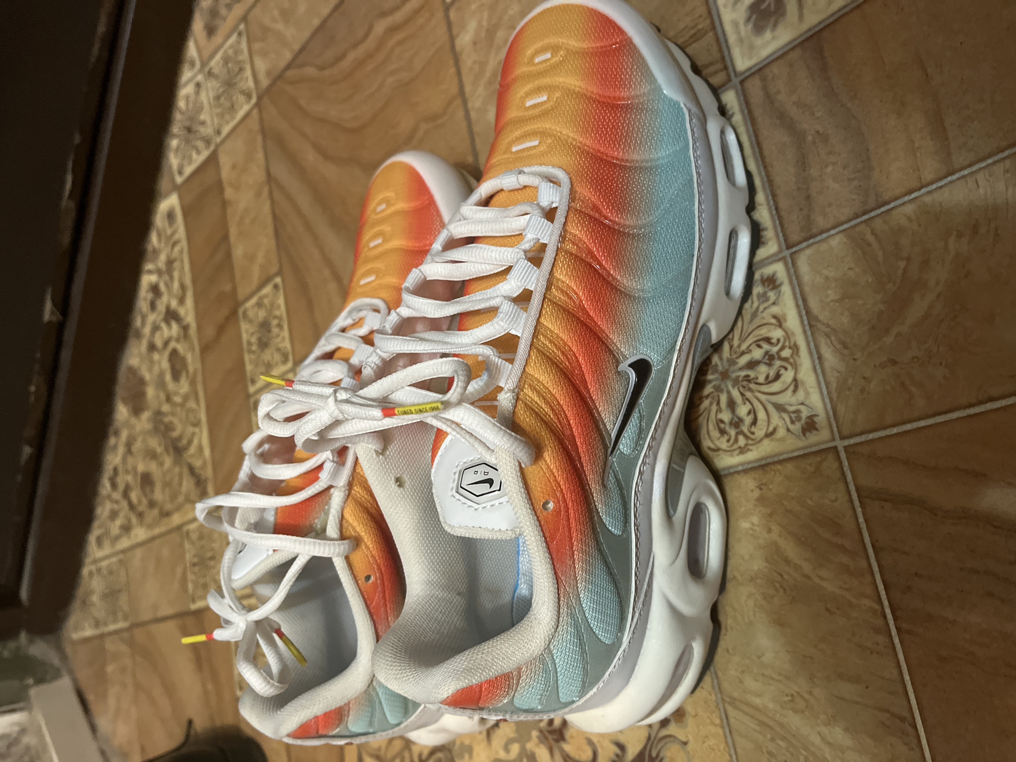 Two Nike Air Max Plus sneakers with a gradient color scheme of teal, orange, and yellow are placed on a floor with tiled sections. The sneakers are positioned side-by-side, with one slightly in front of the other. The background shows a dark wooden surface. There are no people visible in the image. The scene appears to be indoors, likely in a residential setting. There is no information or visual cues in the image that would indicate the location to be in the city of Minsk, Belarus.
