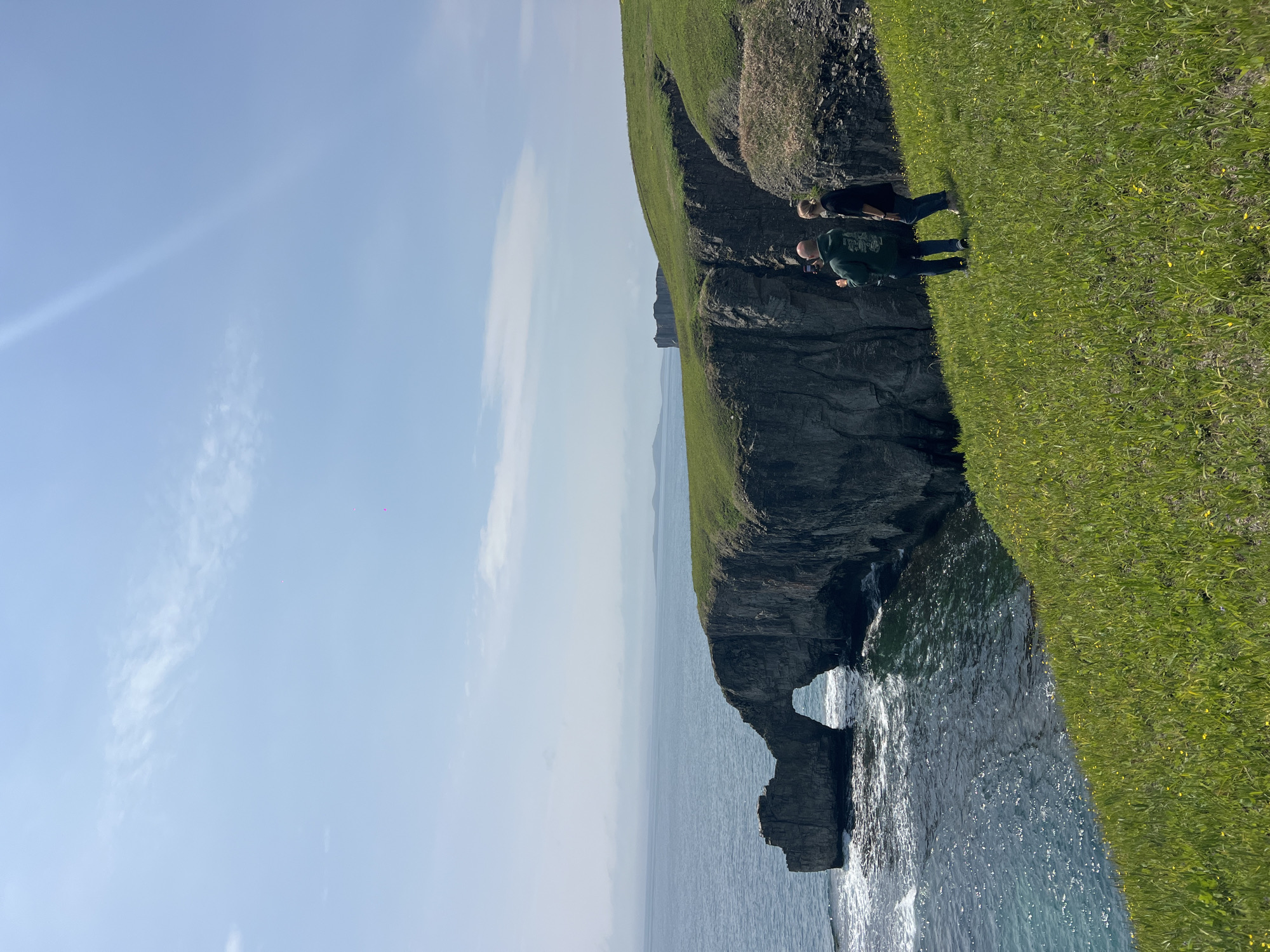 The image depicts a coastal scene with a rocky cliff face and grassy terrain at the top. Two people are standing on the grassy edge, seemingly observing the landscape. The cliff has a natural arch formation at its base, where the water from the ocean flows through. The sky is blue with some wispy clouds. The image does not depict the city of Slavyanka, Russia.