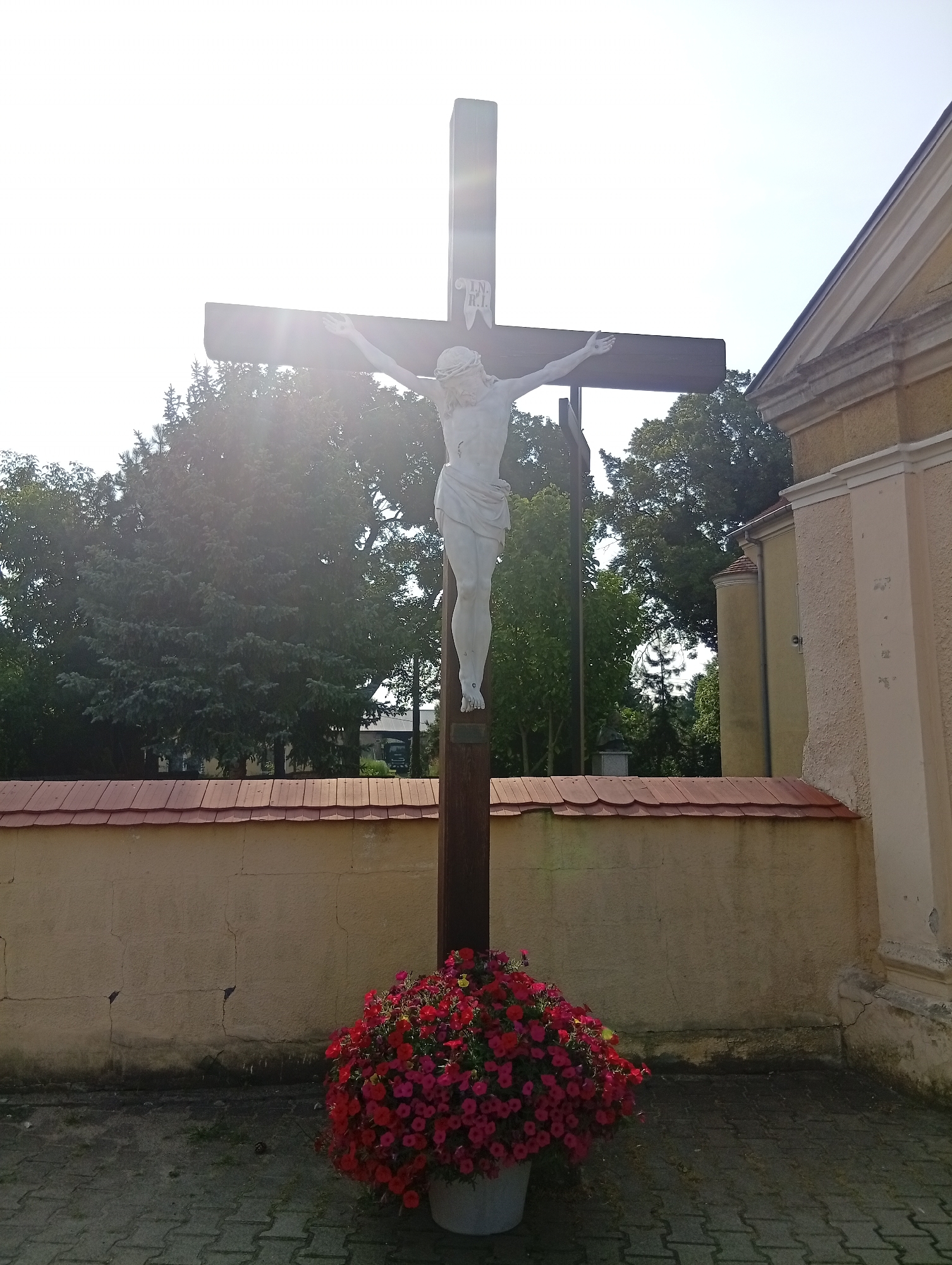 An outdoor statue of Jesus on the cross stands in front of a building and a wall. The statue is made of white material and depicts Jesus with his arms outstretched on a dark wooden cross. Above Jesus' head, on the cross, is a white plaque with the letters "I.N.R.I." in dark lettering. A large planter overflowing with vibrant red flowers is positioned at the base of the cross. Behind the statue, a light-colored building with a red tiled roof is visible to the right. A cream-colored wall with a red tiled coping runs horizontally across the middle ground. Mature trees with dense green foliage form the background, and the sky is bright and overcast. The ground in front of the statue is paved with grey interlocking bricks. The image is taken from a low angle, looking up towards the statue and the sky. This is located in Brzezia Łąka, Poland.