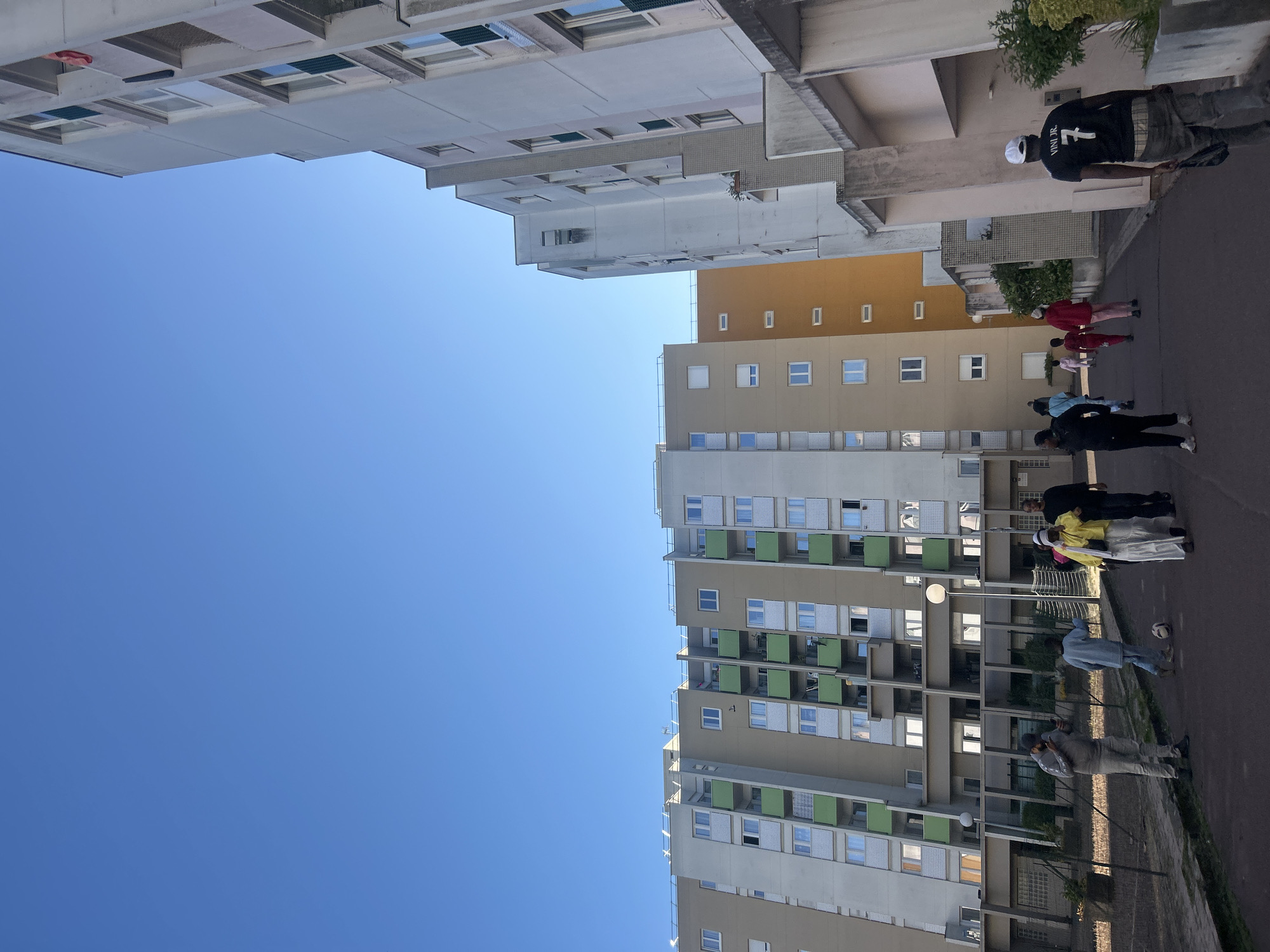 This image captures an urban residential scene in Noisy-le-Grand, France, on a bright, clear day. The primary subjects are several multi-story apartment buildings and a group of people outdoors.
On the left side of the frame, a tall, modern apartment building dominates, characterized by numerous windows and balconies, many of which feature distinctive green panels. In the background and to the right, other apartment blocks are visible, some with beige and orange facades, indicating a large residential complex. The sky is a vibrant, cloudless blue, and strong sunlight casts clear shadows on the buildings and ground, suggesting it is daytime.
Several individuals are present on a paved ground-level area in front of the buildings. In the foreground on the right, a person is seen from behind, wearing a black t-shirt with the text "VINI JR. 7" prominently displayed on the back, along with a white cap. Further down the path, a group of people, including some in red and pink attire, are gathered. Another individual in a light blue top is standing near a black and white soccer ball, suggesting a casual, recreational atmosphere. Other figures are scattered throughout the scene, either standing or walking along the path. Green foliage and bushes are visible near the bases of some buildings. The overall impression is a typical day in a residential neighborhood.