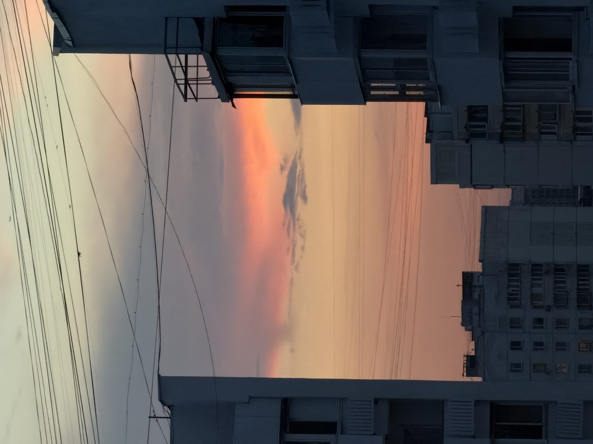 This is a vertical, full-shot, outdoor photograph taken at twilight in Khimki, Russia. The image is dominated by the sky, which transitions from pale yellow at the horizon to a soft lavender and gray at the top. There are multiple black power lines crisscrossing the sky. In the mid-ground and foreground, several apartment buildings are visible, rendered in shades of gray and white. The buildings are utilitarian in design, featuring numerous windows and balconies. One building on the right has a distinctive balcony with a metal railing. The sky exhibits a dramatic sunset with vibrant pink and orange clouds concentrated in the upper half of the frame. No people are visible in the image. The overall atmosphere is calm and somewhat desolate, characteristic of an urban twilight scene.