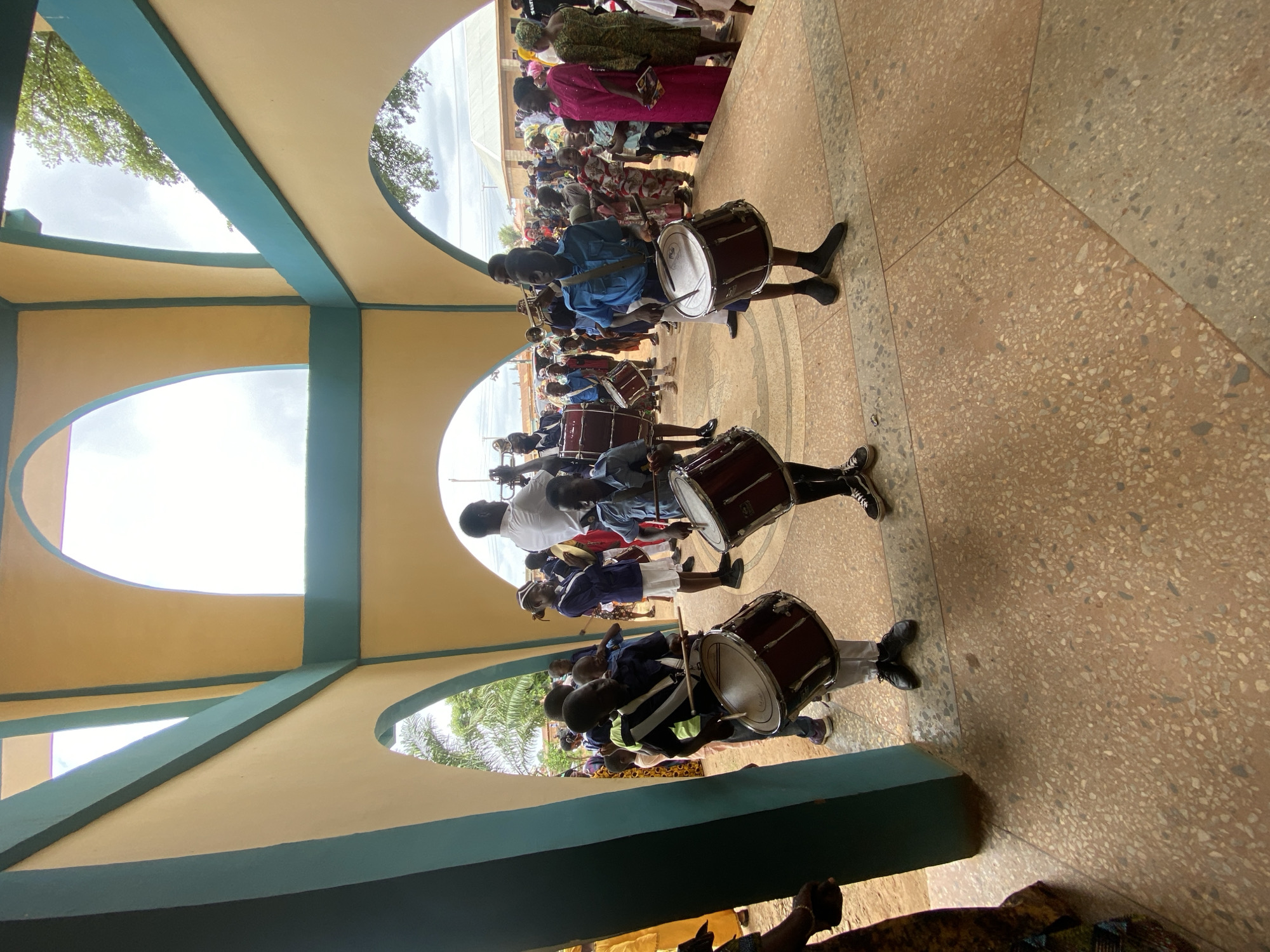 Captured from a low-angle perspective inside a building in Kafanchan B, Nigeria, the image features a line of drummers in a band. The drummers, all dark-skinned, are dressed in assorted uniforms and are carrying and playing large drums. They are arranged in a line, moving right to left, with an audience visible in the background. The building's architecture includes arched openings that frame the scene, with cream-colored walls and blue-green accents. The floor is a terrazzo style, made up of marble chips.