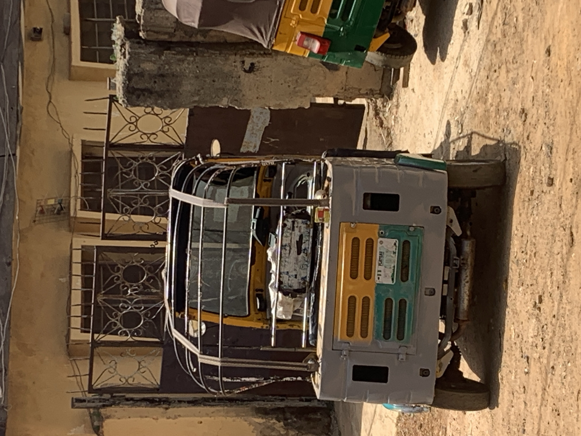 A three-wheeled vehicle with a metal cage around the passenger compartment is parked next to a building. The vehicle's rear panel is painted grey, with yellow and green vertical stripes. The building has windows with decorative metal grilles. Another three-wheeled vehicle is partially visible in the background. The location is Rafin Par, Nigeria.
