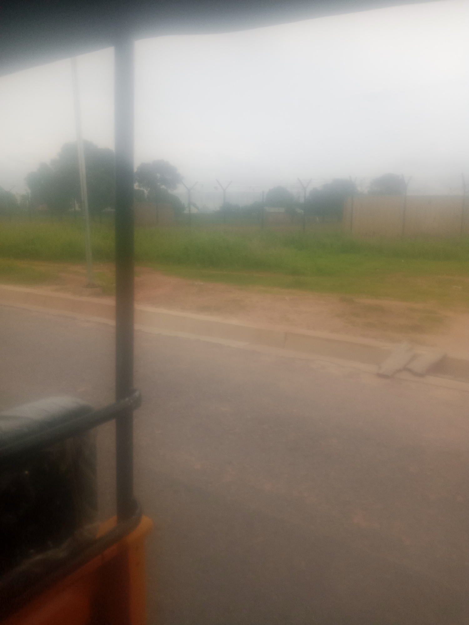 The image captures a hazy scene from the perspective of a moving vehicle in Jalingo, Nigeria. In the foreground, a dark metal bar runs horizontally across the bottom, with a dark object to its left and an orange structure below. A vertical dark pole divides the frame in the center. The road is visible in the lower half of the image, with a concrete curb bordering a patch of green grass and dirt. In the background, trees are silhouetted against a bright, foggy sky. A chain-link fence with metal posts stretches across the mid-ground, suggesting a perimeter or boundary. The overall atmosphere is obscured by fog or haze, contributing to a muted color palette and soft focus.