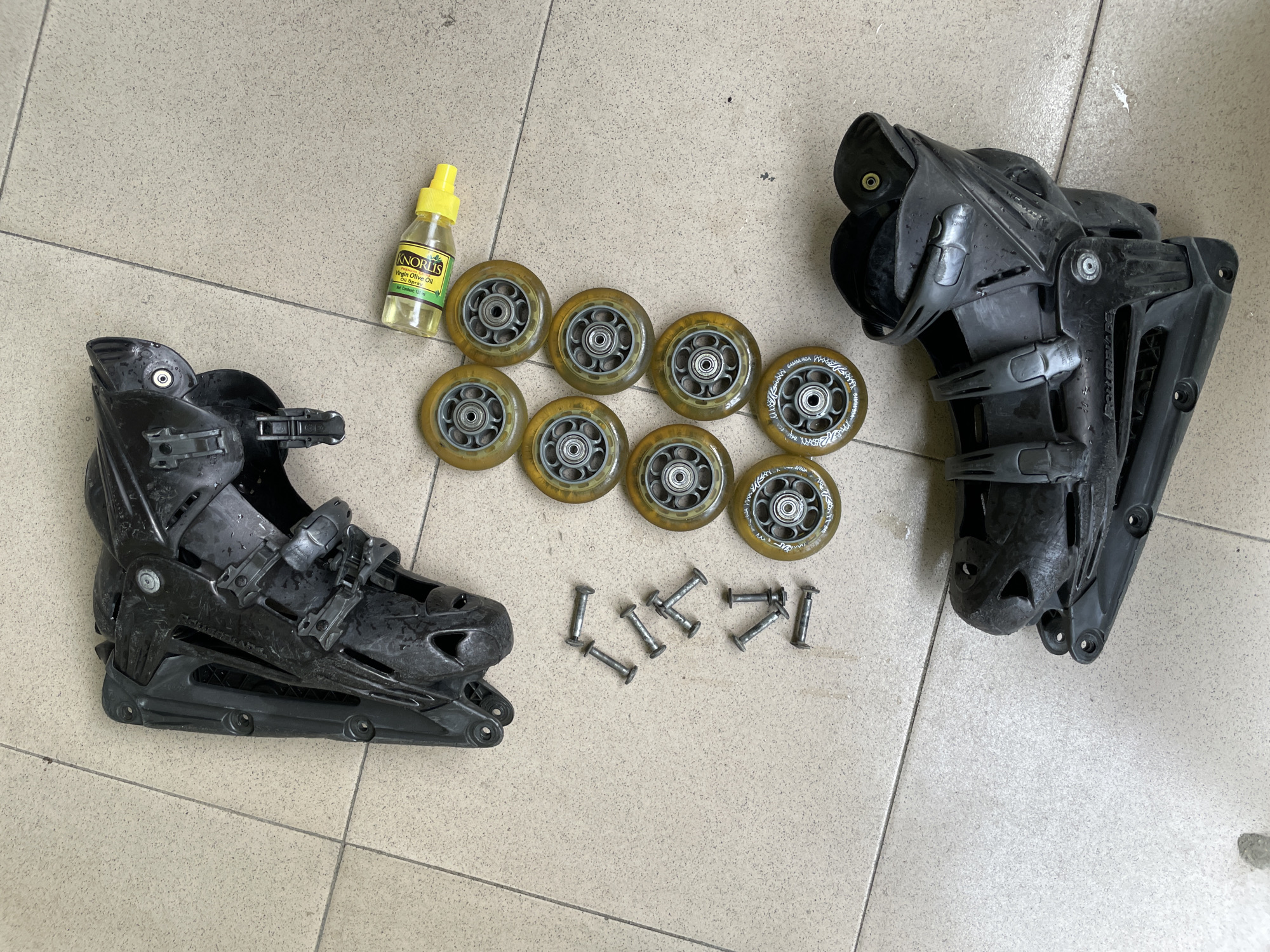 A pair of disassembled inline skates, including two black boot shells, eight yellow wheels, and ten silver bolts, are laid out on a tiled floor. A small yellow bottle of lubricating oil is also present. The scene appears to be indoors, possibly in a workshop or storage area, with the light casting shadows on the floor. The overall impression is one of maintenance or preparation for repair. The location is a tiled floor in the city of Ado, Nigeria.