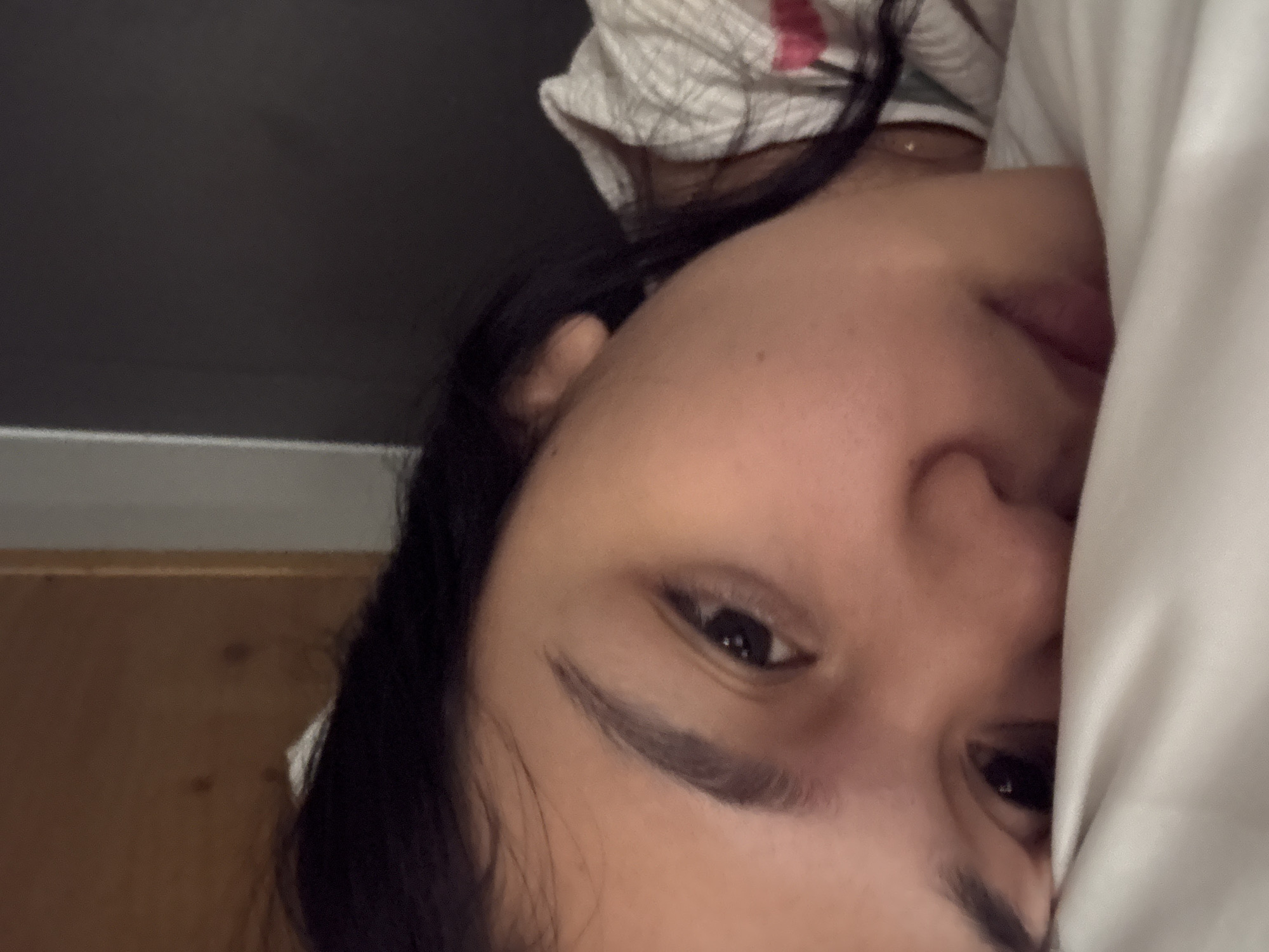 The image depicts a close-up view of an individual, stated to be located in Davao City, Philippines, lying down in an indoor setting. The subject, a young adult with dark hair and prominent dark eyebrows, is positioned with their head resting on a white pillow or bedding, which occupies the lower half of the frame. Their face is angled towards the camera, with the right eye fully visible and the left eye partially obscured by the bedding. The expression is subdued, appearing either sleepy or pensive.
The individual is wearing a light-colored, patterned top, featuring subtle textures and hints of pink or red within its design. A small, reflective, possibly gold-toned stud or similar adornment is visible near the right side of the neck/collarbone area. The background on the upper left consists of a light brown wooden surface, while the upper right features a plain, dark grey or black wall, with a faint lighter vertical line suggesting a structural corner or frame. The overall lighting conditions are dim, consistent with an indoor environment such as a bedroom.